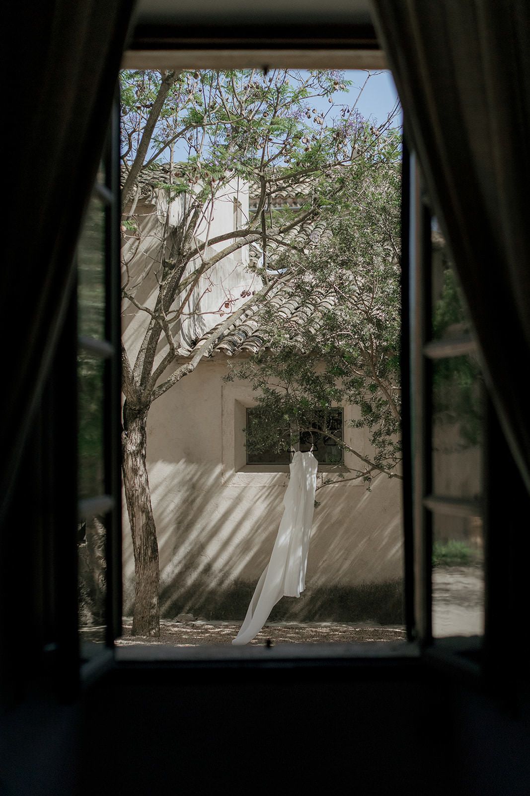 White curtains blow in the breeze through an open window frame overlooking a sunny courtyard with trees and walls.