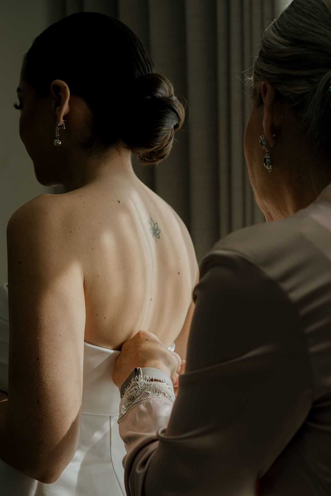 Bridesmaid carefully fastening buttons on back of elegant white bridal gown before wedding ceremony.