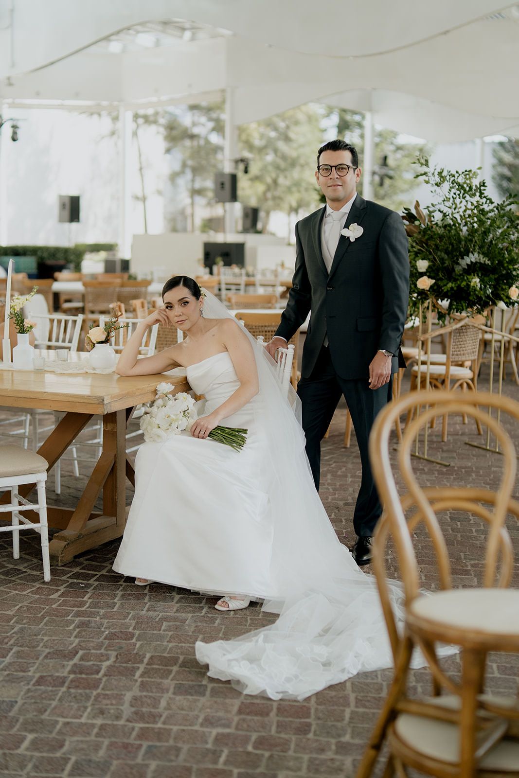 Bride in strapless white gown sits at wooden table beside groom in dark suit at elegant outdoor wedding reception.