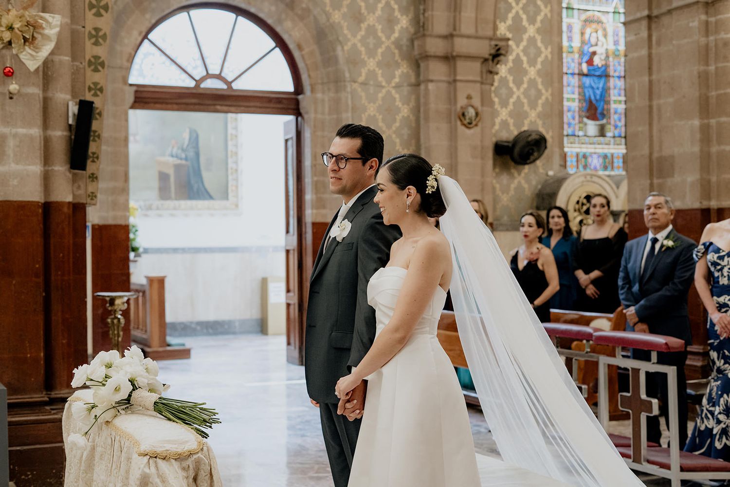 Bride and groom holding hands at the altar during a church wedding ceremony with guests seated in pews.