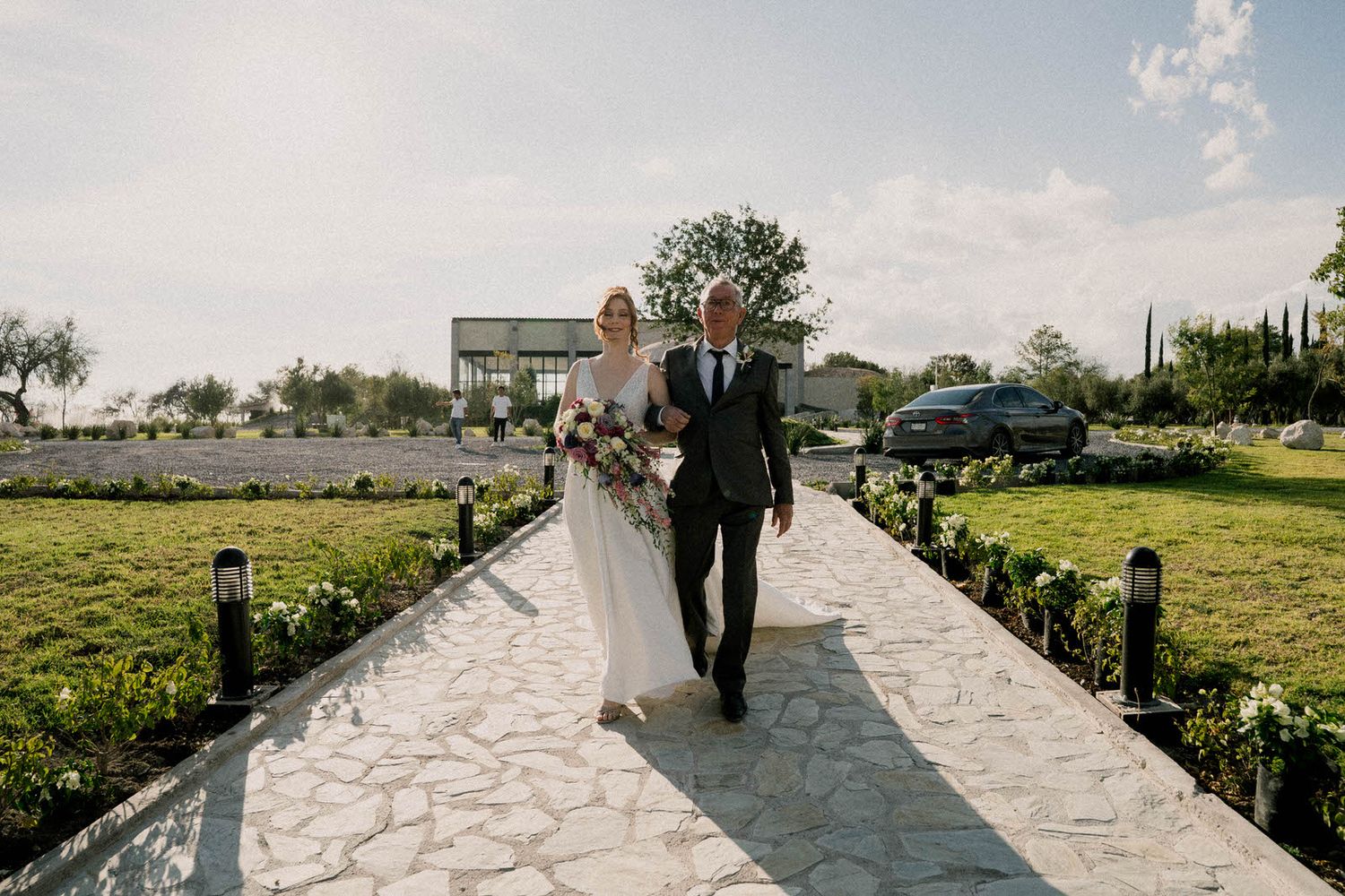 Outdoor wedding venue with stone pathway leading to wooden pergola surrounded by green landscaping.