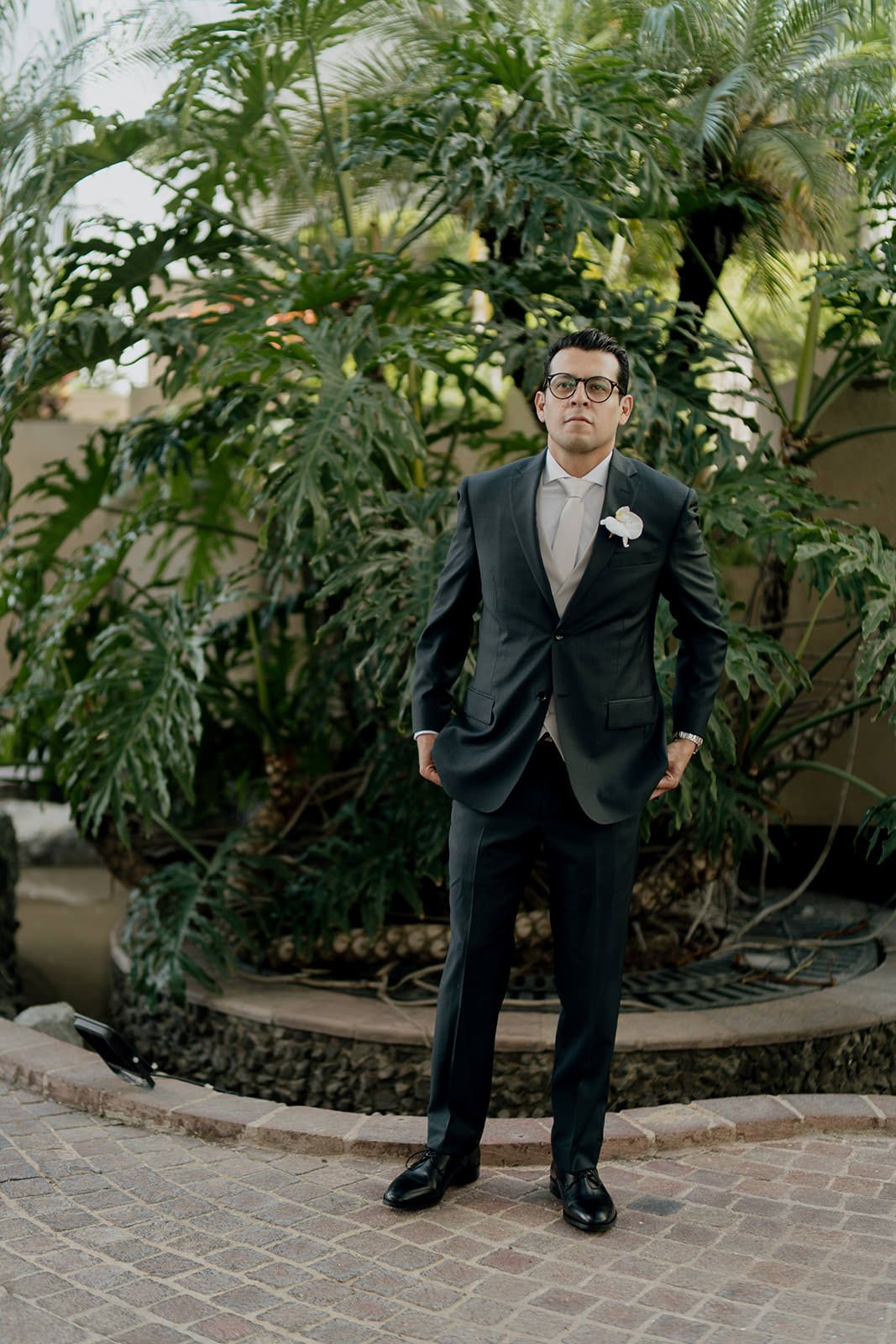 Dapper groom in dark suit with boutonniere stands confidently among tropical palm plants outdoors.