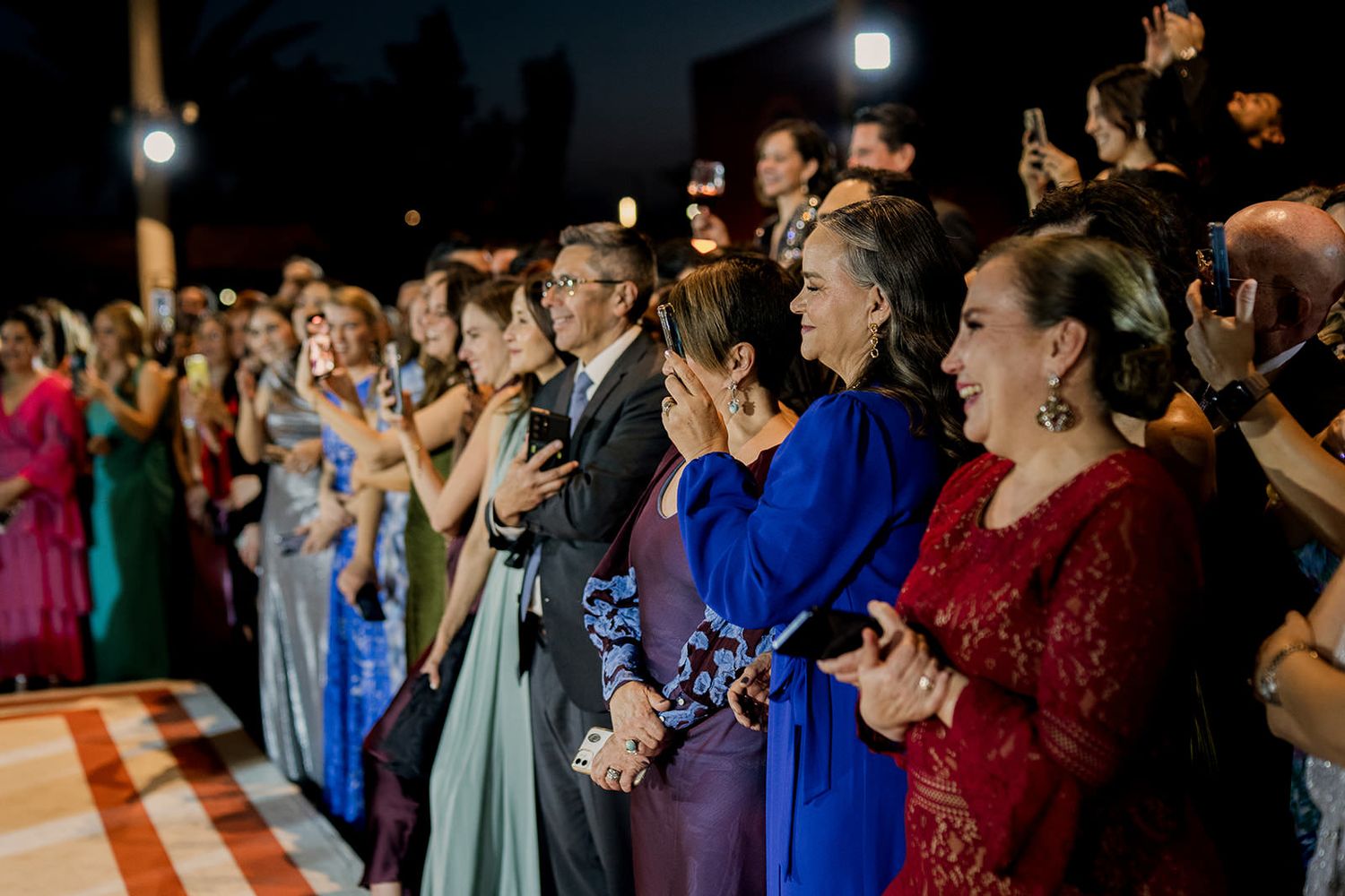 Elegantly dressed wedding guests clapping and celebrating at an outdoor evening reception.