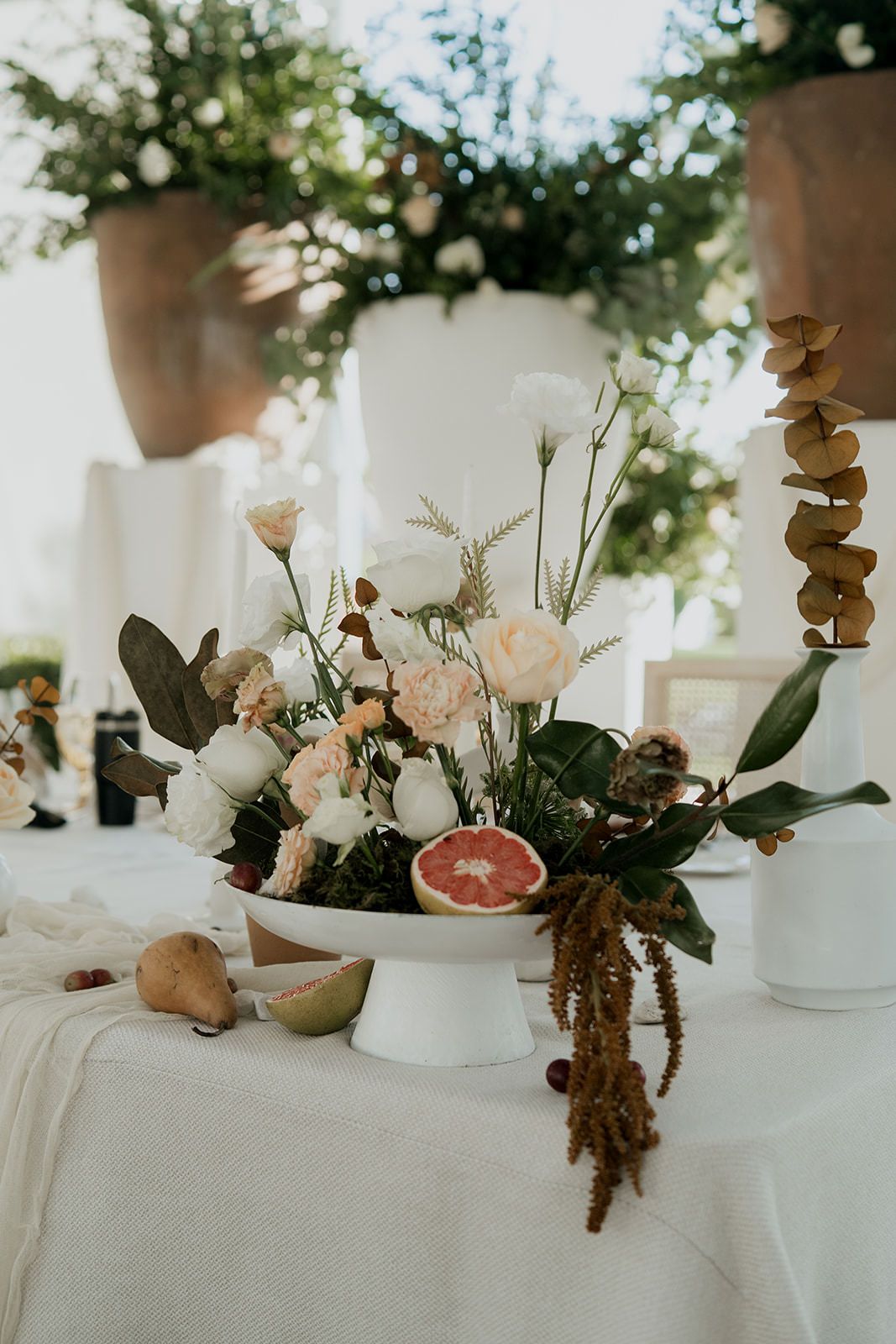 Elegant wedding centerpiece with white and peach roses, greenery, and a halved grapefruit on a white pedestal dish.