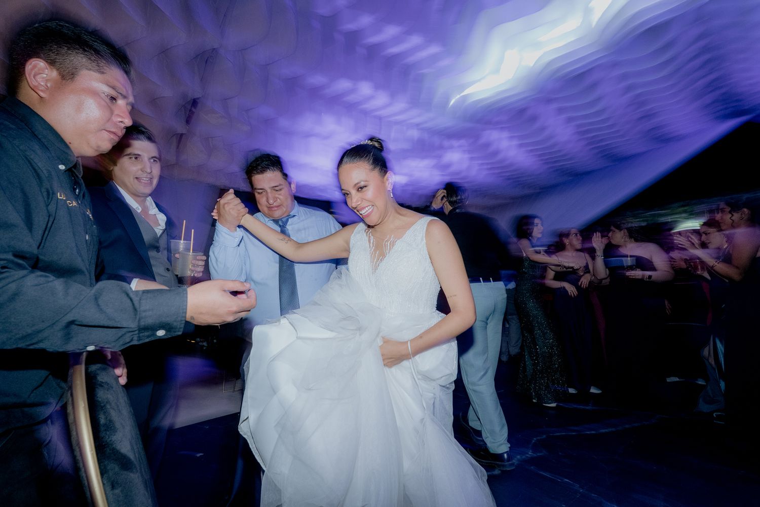 Bride in white gown dancing with guests at a wedding reception under blue lights.
