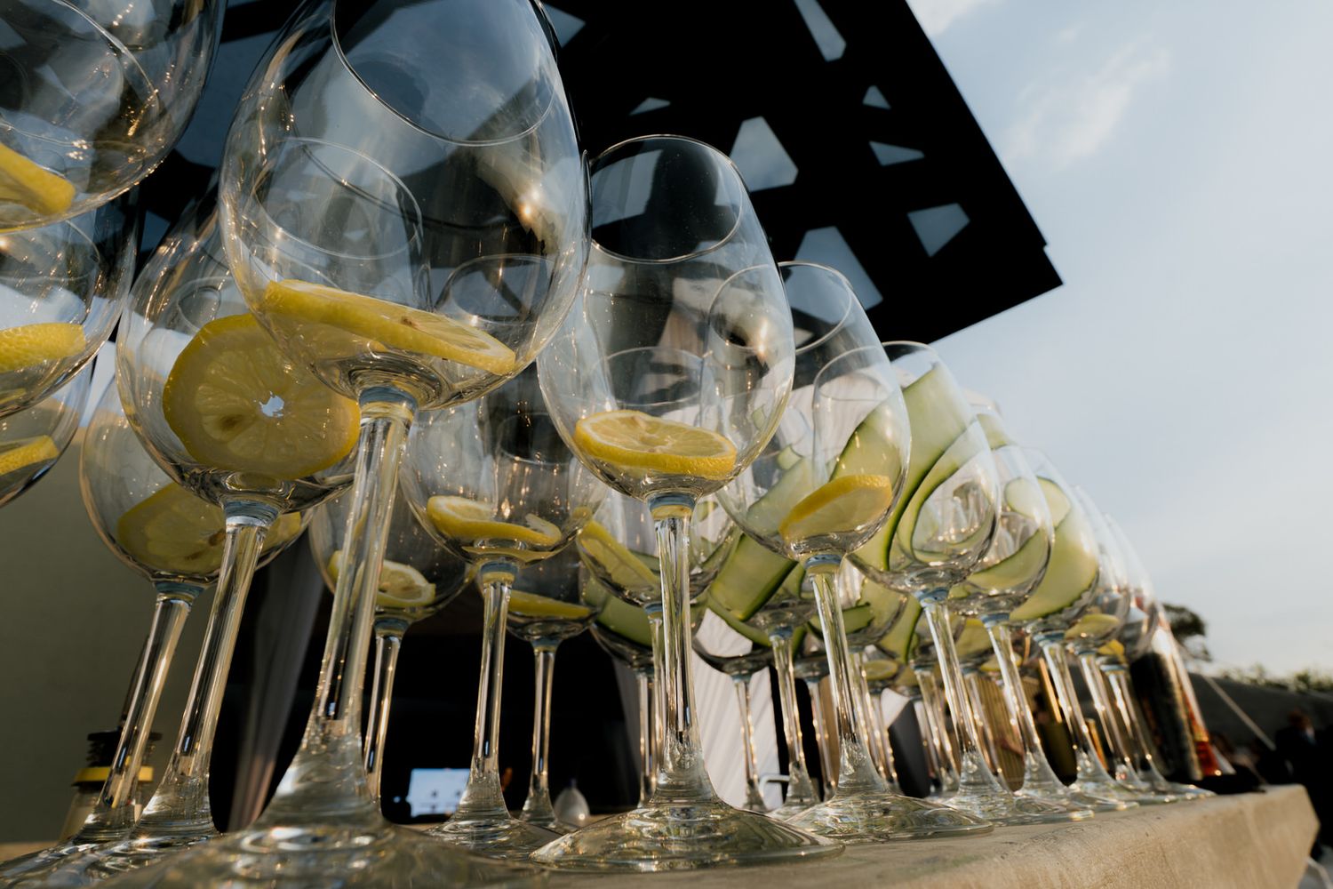 Row of wine glasses filled with lemon slices lined up on outdoor bar table at wedding reception.