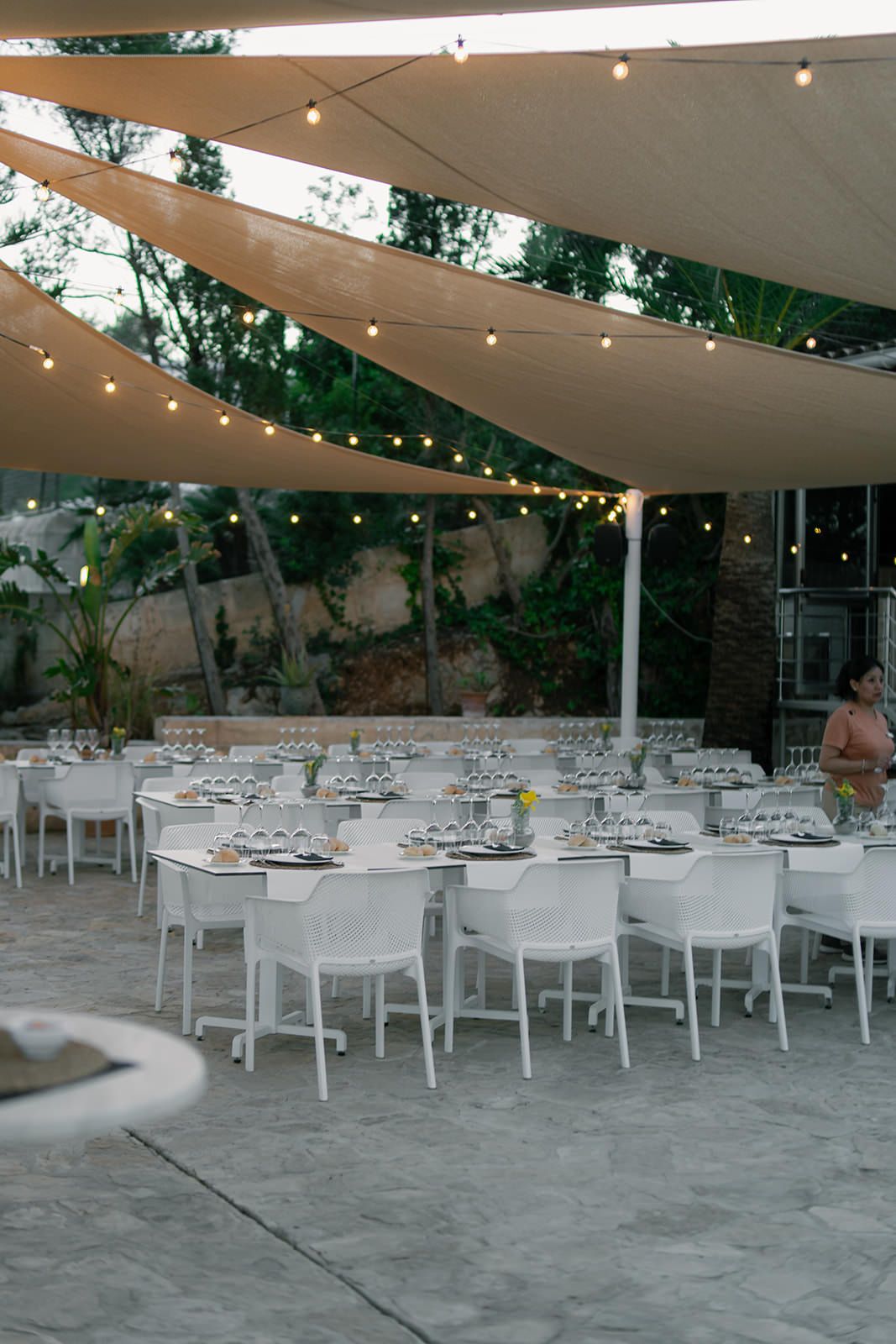 White dining tables and chairs arranged under a canopy with string lights for an elegant outdoor reception.