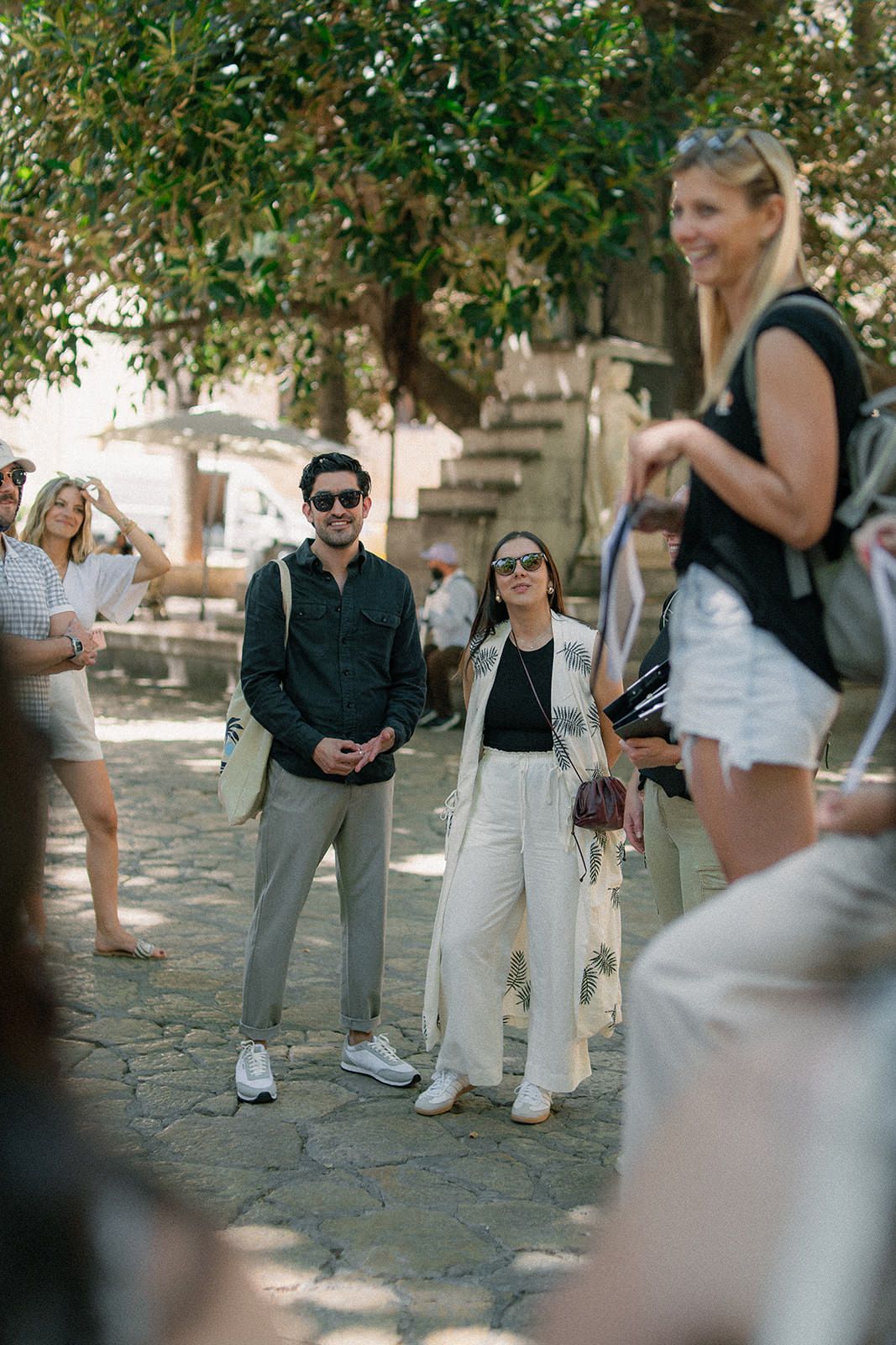 A group of people gathered outdoors on a stone paved area beneath trees on a sunny day.