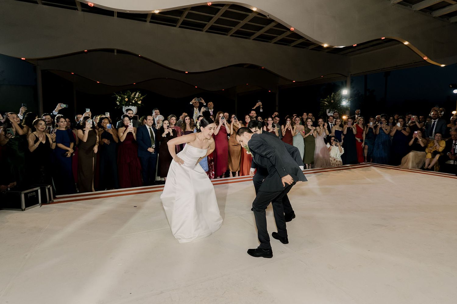 Groom dips bride on white dance floor surrounded by cheering wedding guests at night.