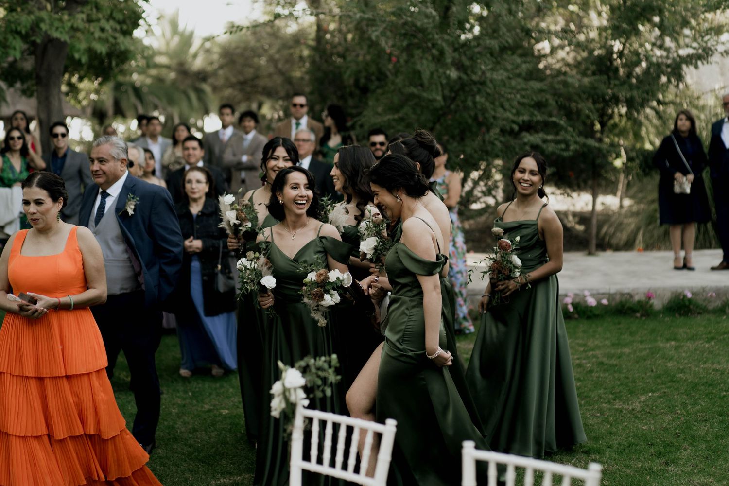 Wedding attendees in colorful formal wear gather around white chairs during outdoor ceremony.