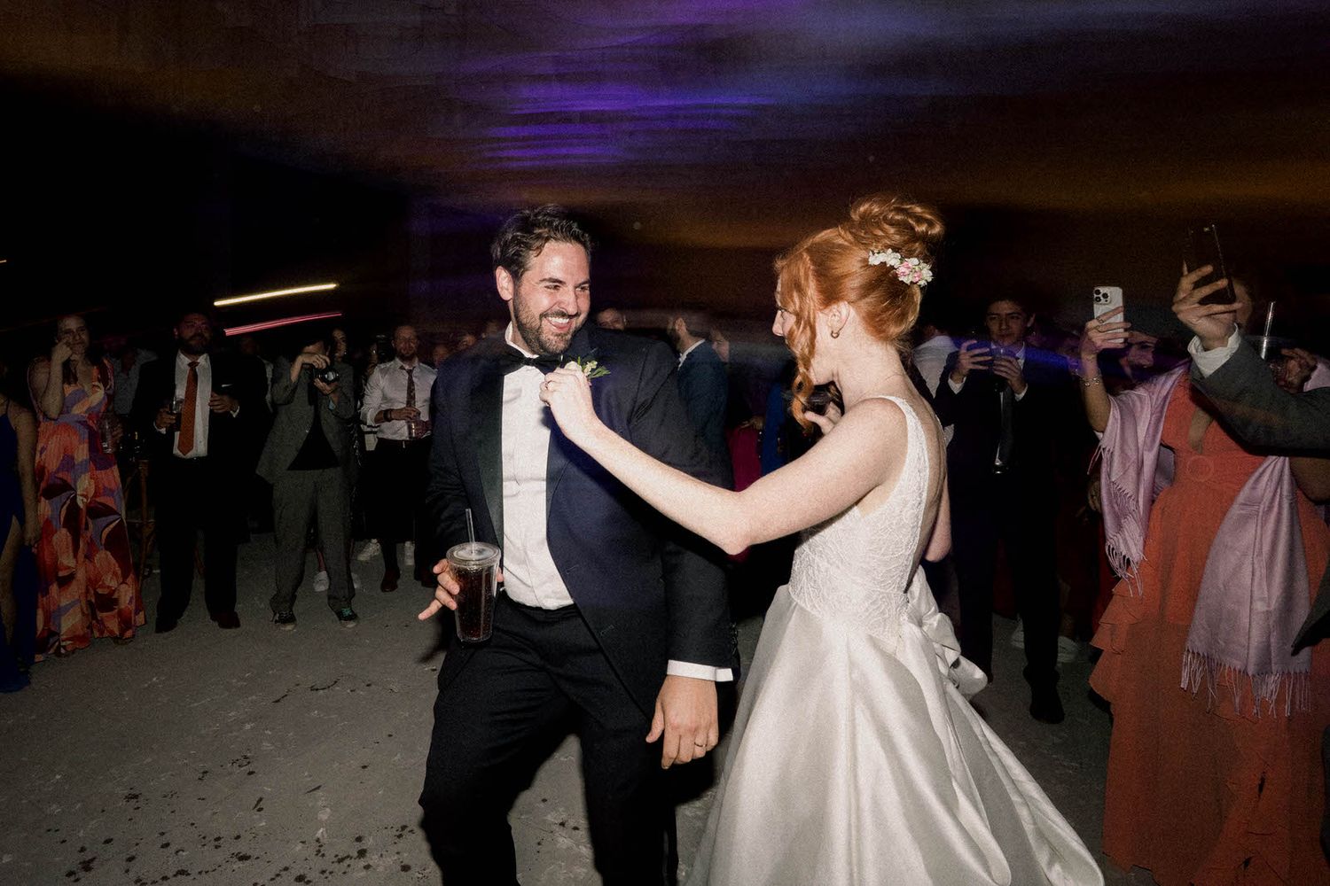 A couple dances together at their wedding reception under dramatic lighting with guests watching in the background.