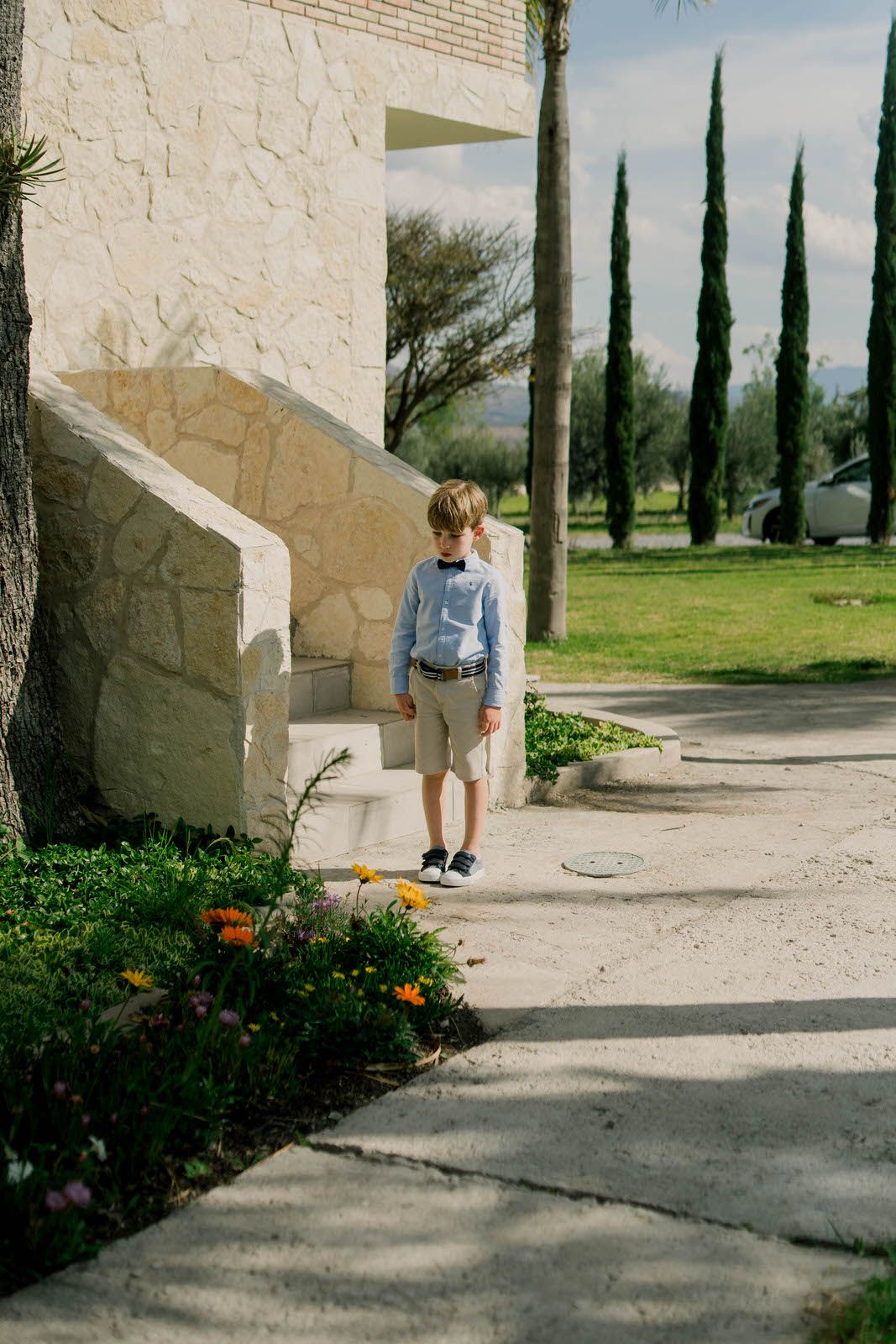 A young ring bearer in blue shirt and khaki shorts walks along a stone path lined with cypress trees.