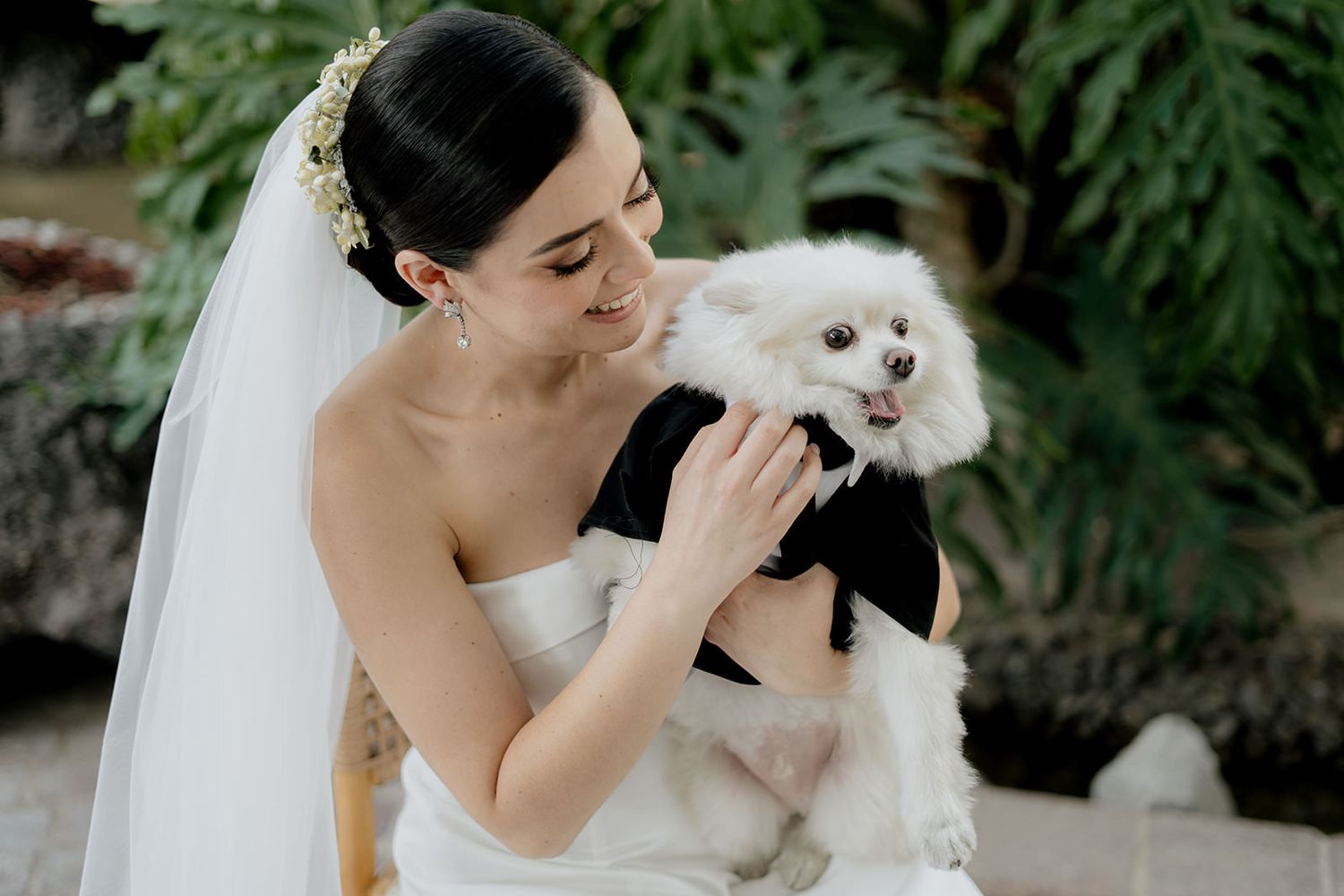 Radiant bride in strapless white gown and veil lovingly holds a fluffy white dog outdoors.
