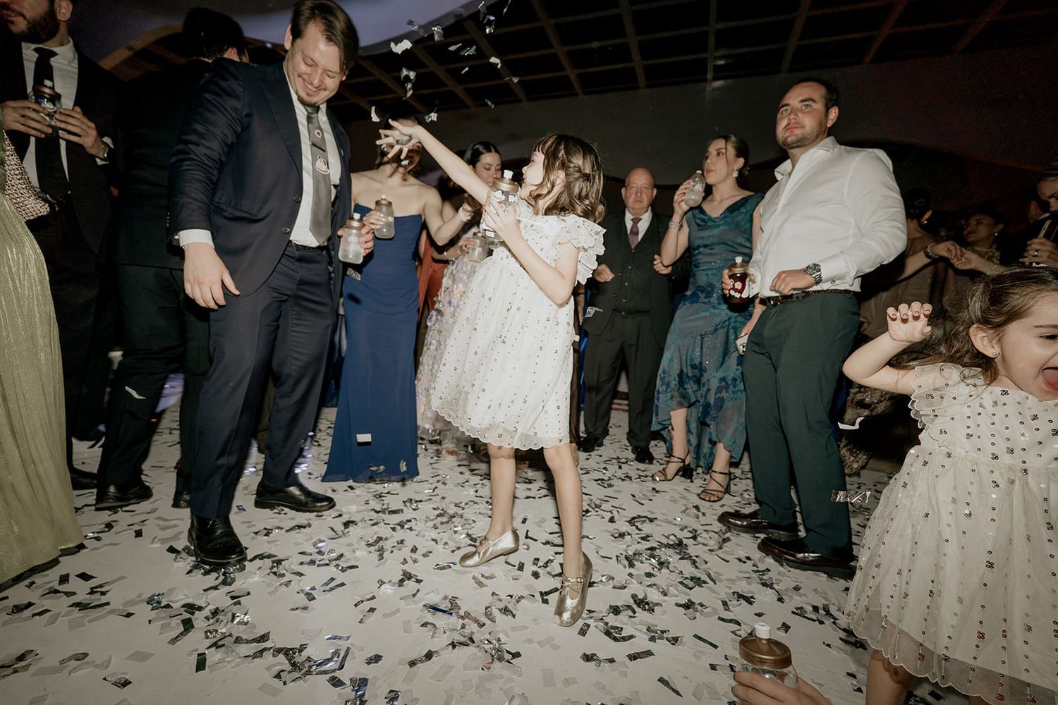 Young flower girl dances joyfully in confetti with wedding guests on decorated reception dance floor at night.