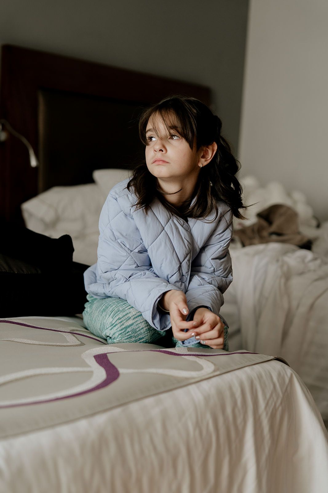 Young girl in blue quilted jacket sitting cross-legged on hotel bed during wedding day preparations.