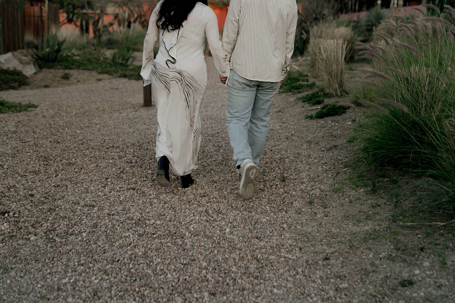 Wedding guests walk along a gravel path lined with desert plants at dusk.