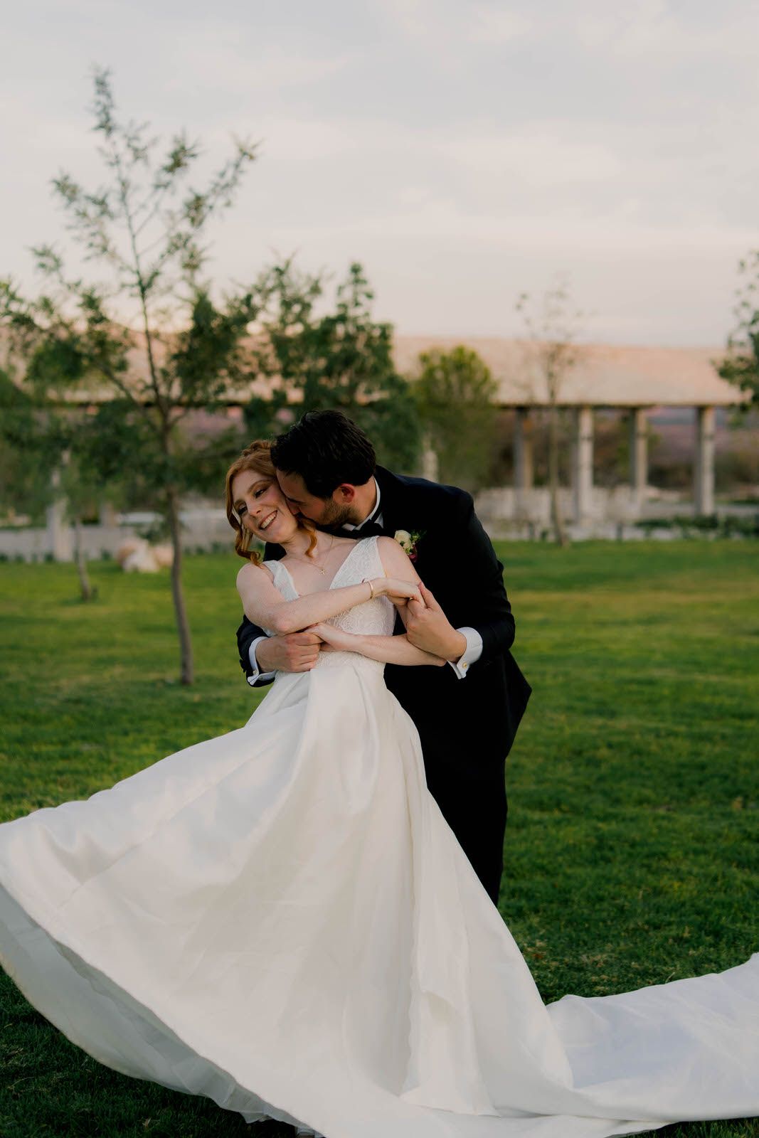 A wedding couple dances playfully on green grass during golden hour with the bride's dress flowing.