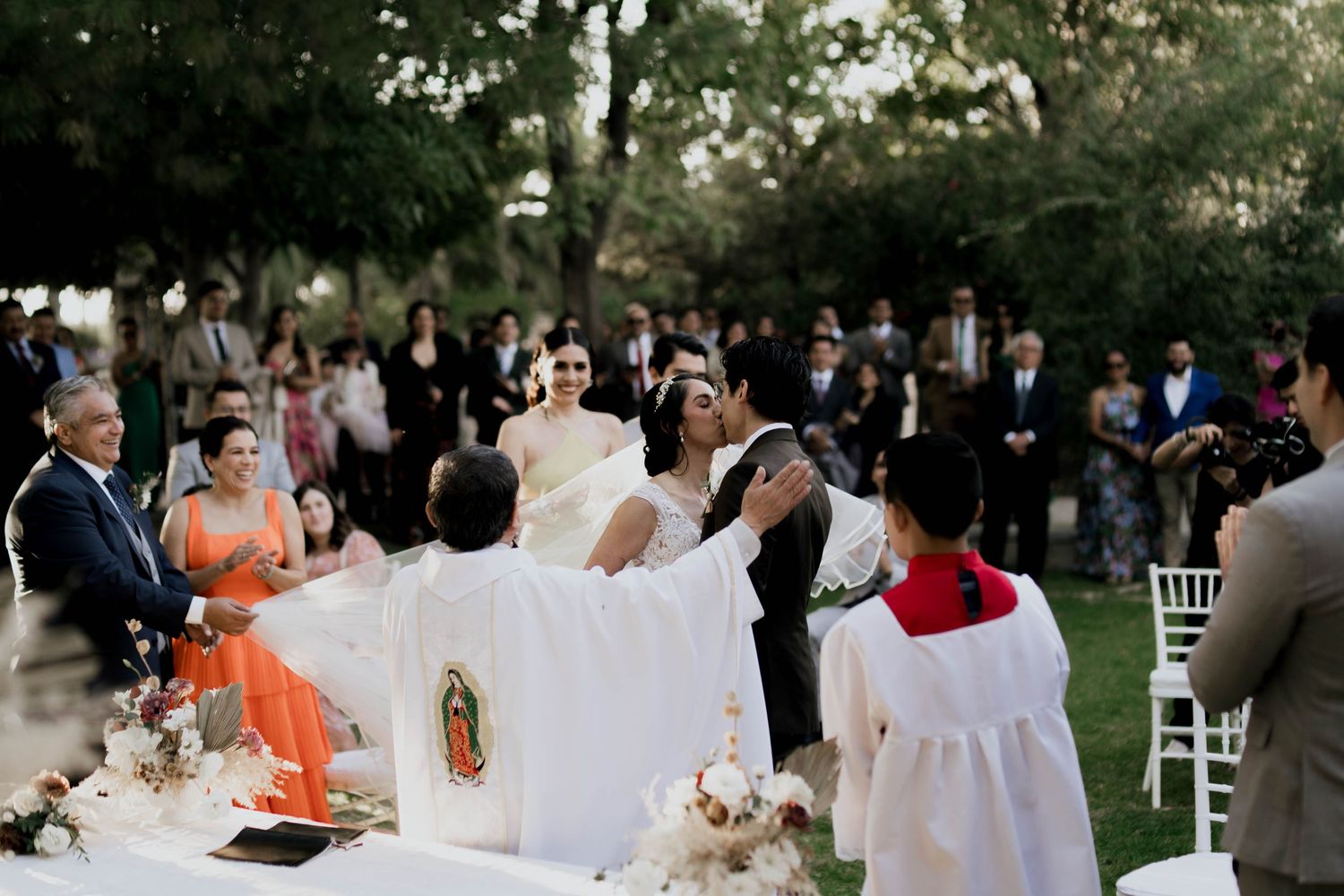 Wedding celebration scene with guests and white decor under trees at an outdoor venue.