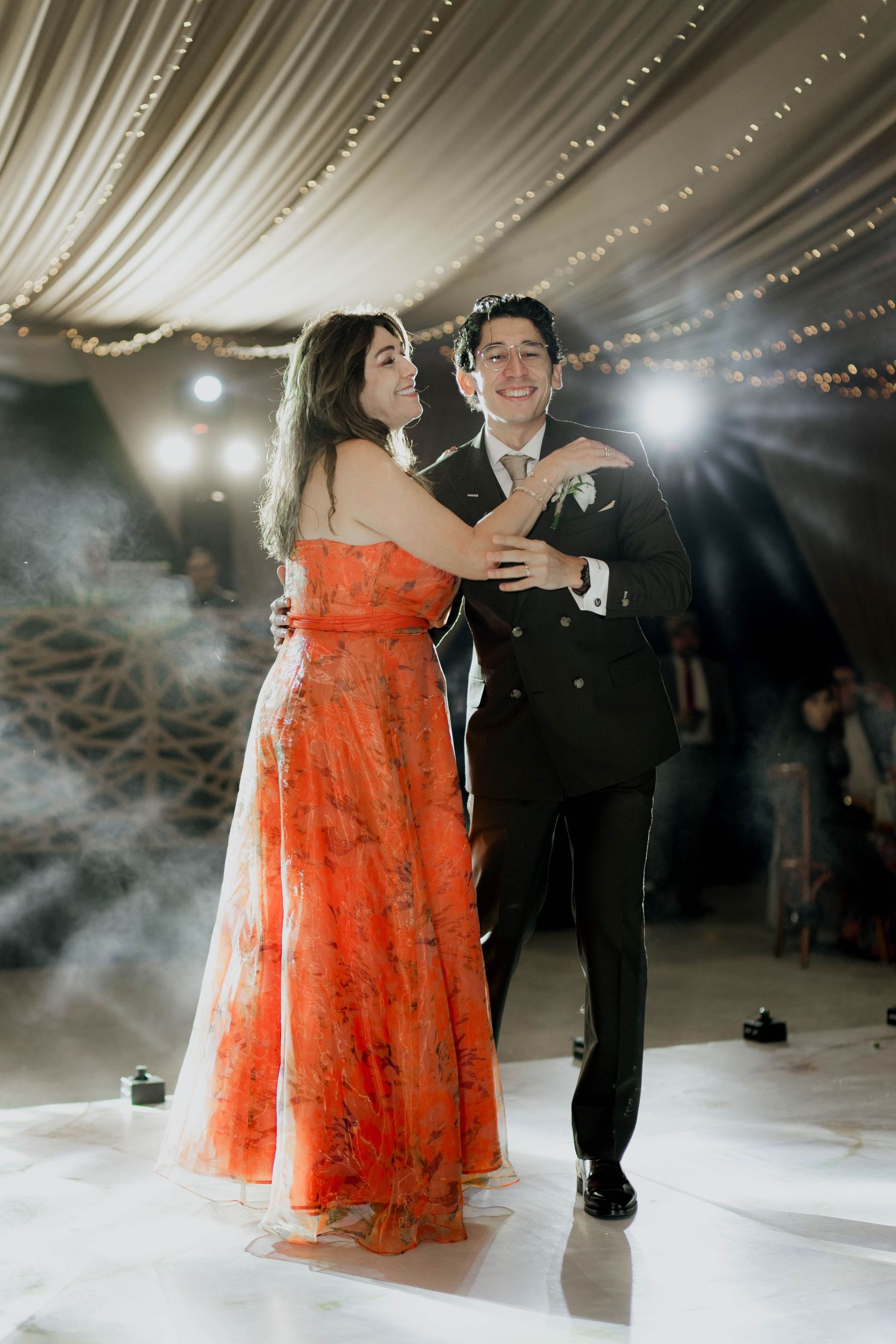 Couple dancing under string lights with smoke effects at wedding reception.