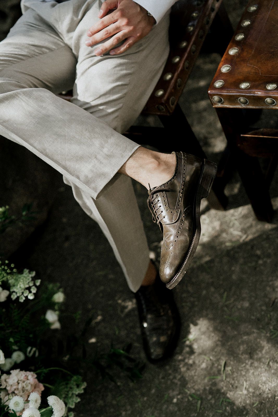 Close-up of polished brown leather dress shoes worn with light gray suit pants on a stone surface.