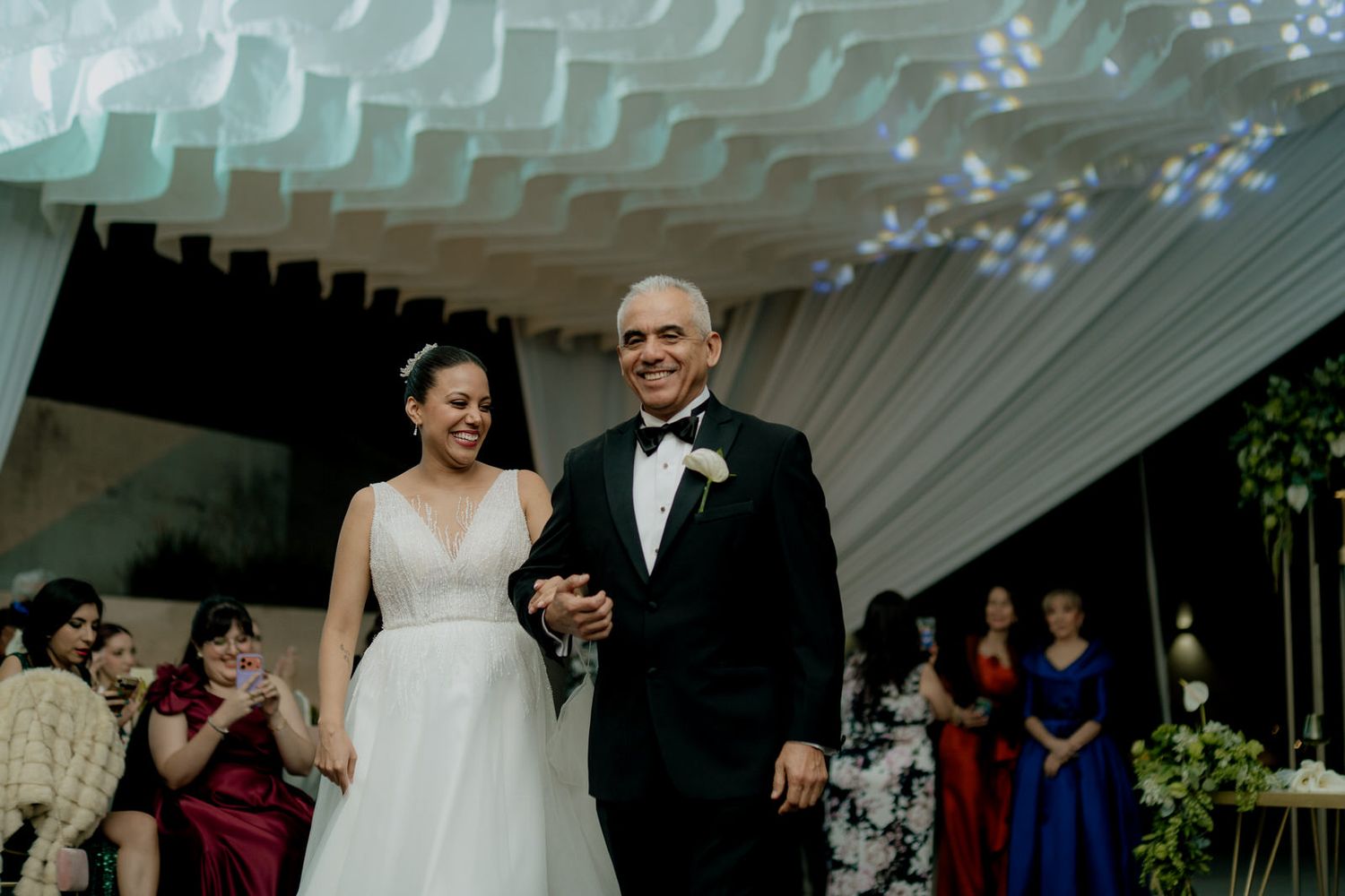A smiling bride in a white gown walks down the aisle with an older man in a tuxedo.