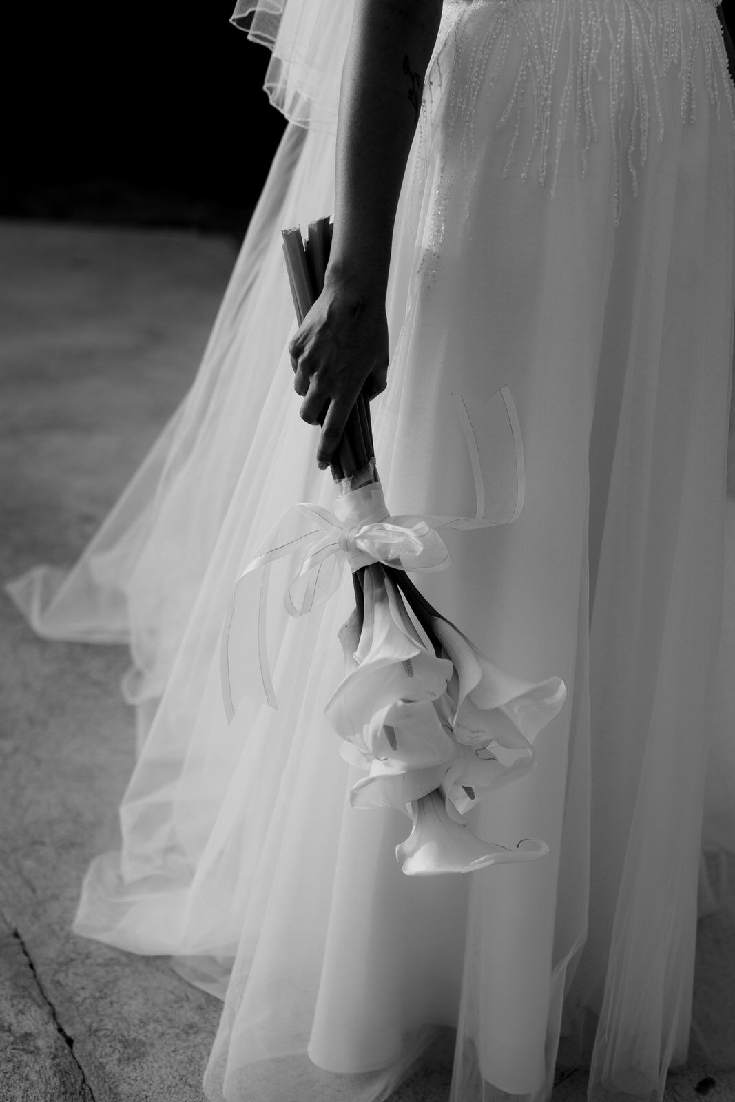 Black and white close-up of bride holding elegant white calla lily bouquet with ribbon against flowing tulle wedding gown.