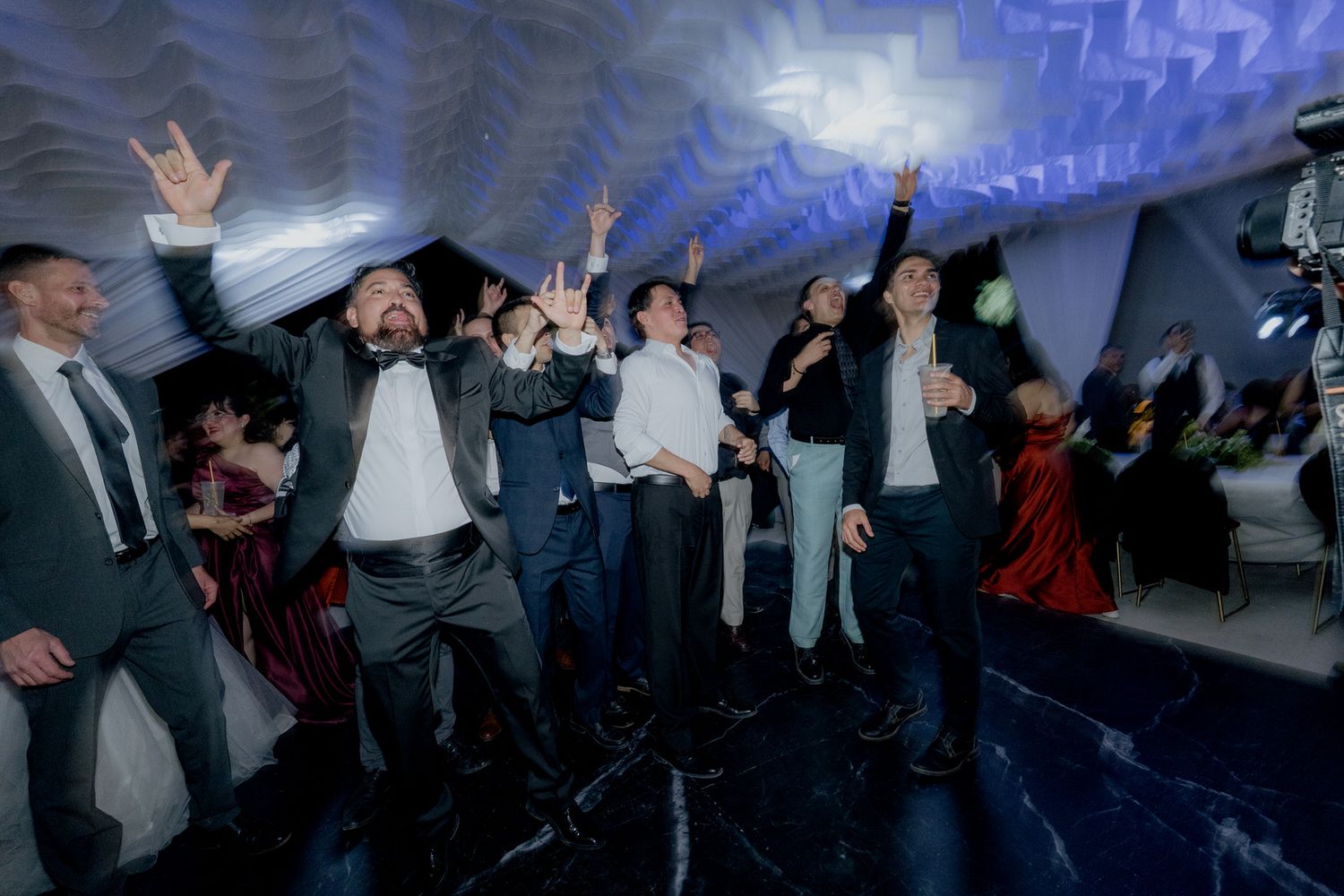 Group of men in suits cheer and raise hands joyfully on a wedding dance floor.