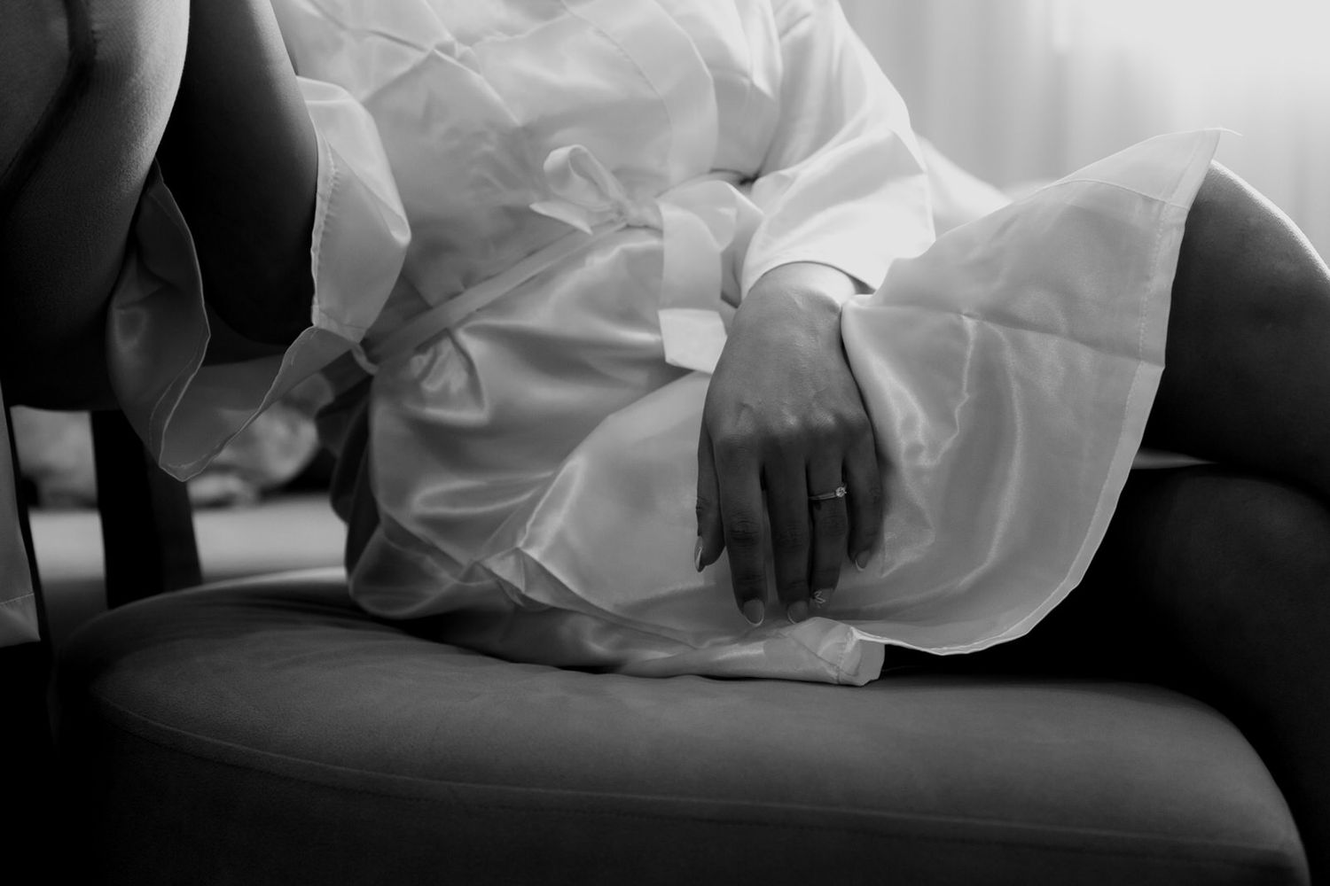 Black and white photo of a bride in white gown sitting gracefully on a chair, hands folded in lap.