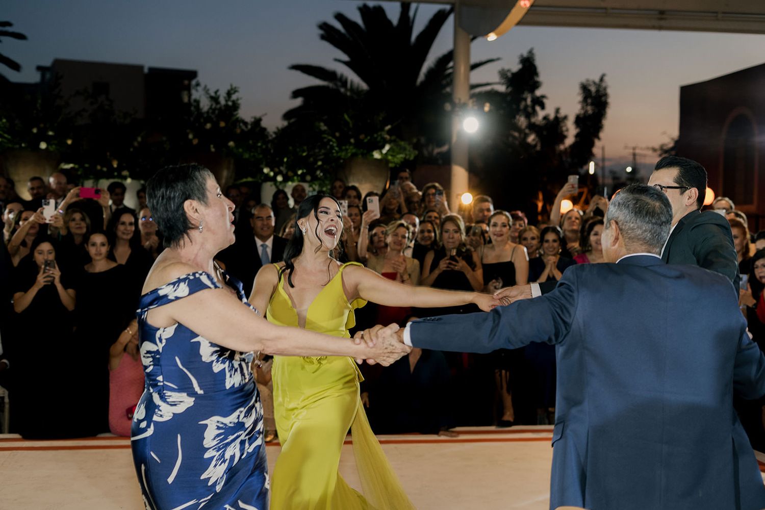 Guests dancing joyfully at an outdoor wedding reception at night, with crowd watching in background.