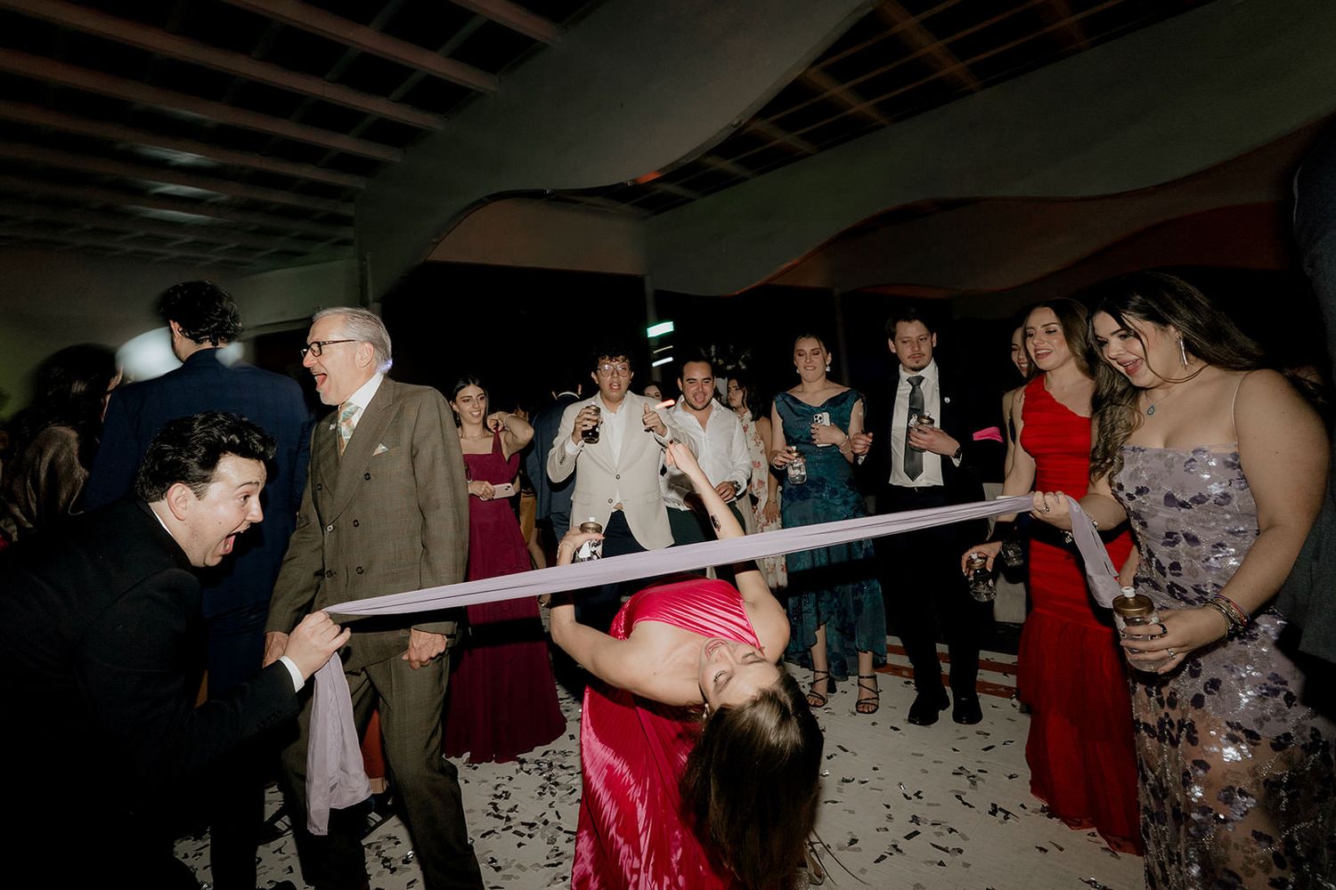 Wedding guests watching a woman do the limbo under a ribbon at a lively reception party.