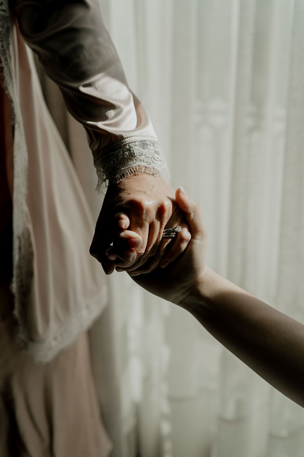 Bride and groom holding hands, showcasing wedding ring and lace bridal sleeve near sheer curtains.