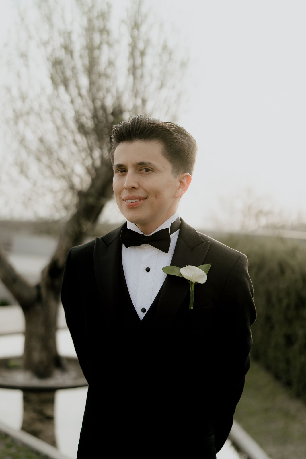 Groom in black tuxedo with white boutonniere and bow tie smiling outdoors at wedding ceremony.