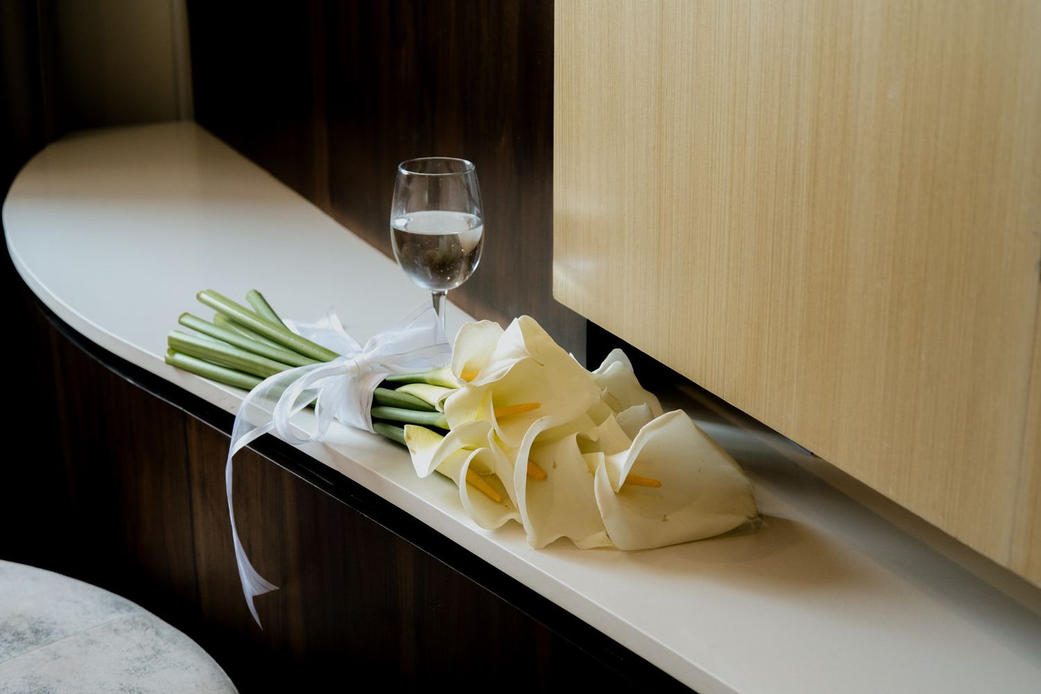 White calla lily wedding bouquet with ribbon beside a glass of water on an elegant shelf.