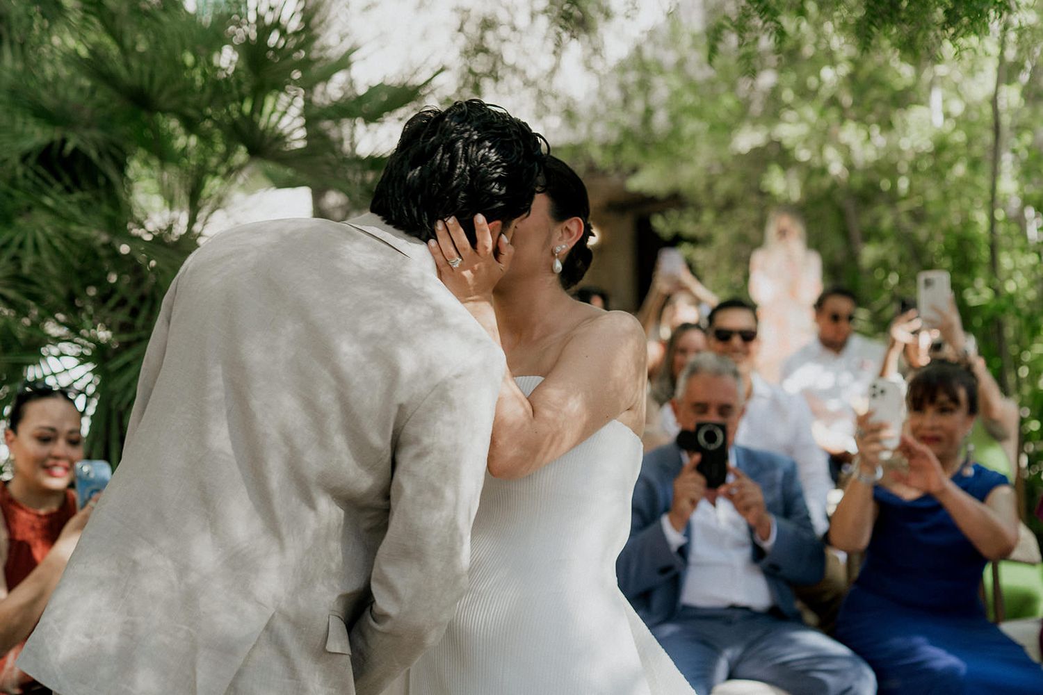 Wedding ceremony guests gather outdoors under trees with white flowing fabric decorations.