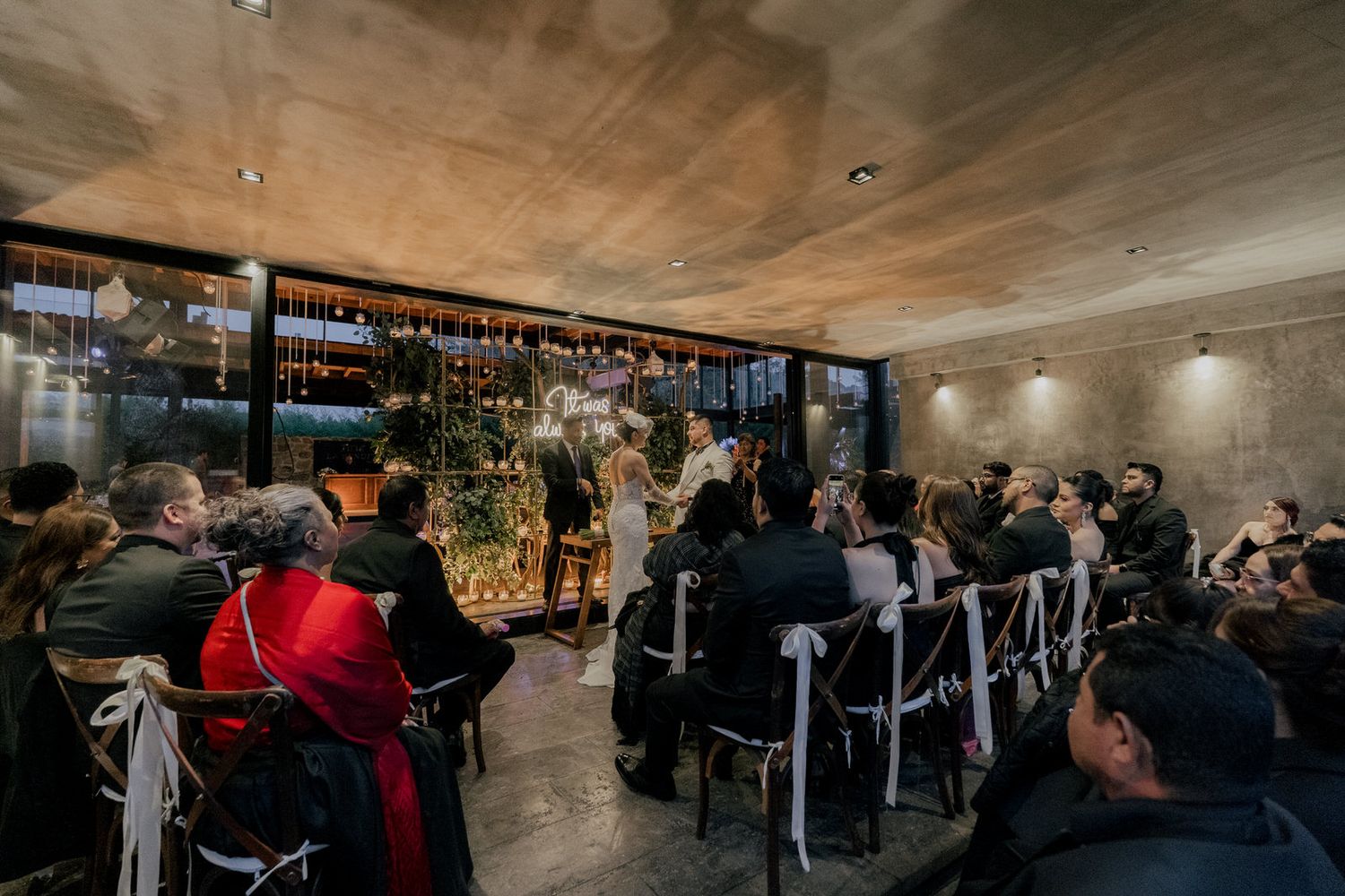 Wedding guests seated at tables in an industrial-style venue with hanging lights and greenery.