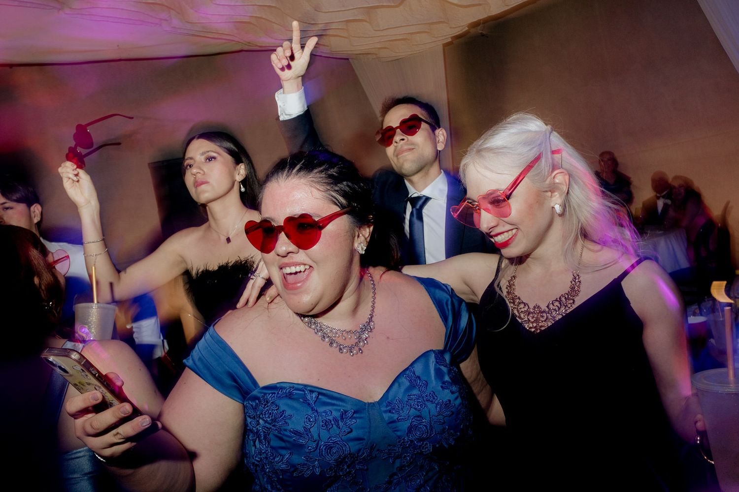 Women wearing red heart-shaped sunglasses dancing and celebrating at a wedding reception.