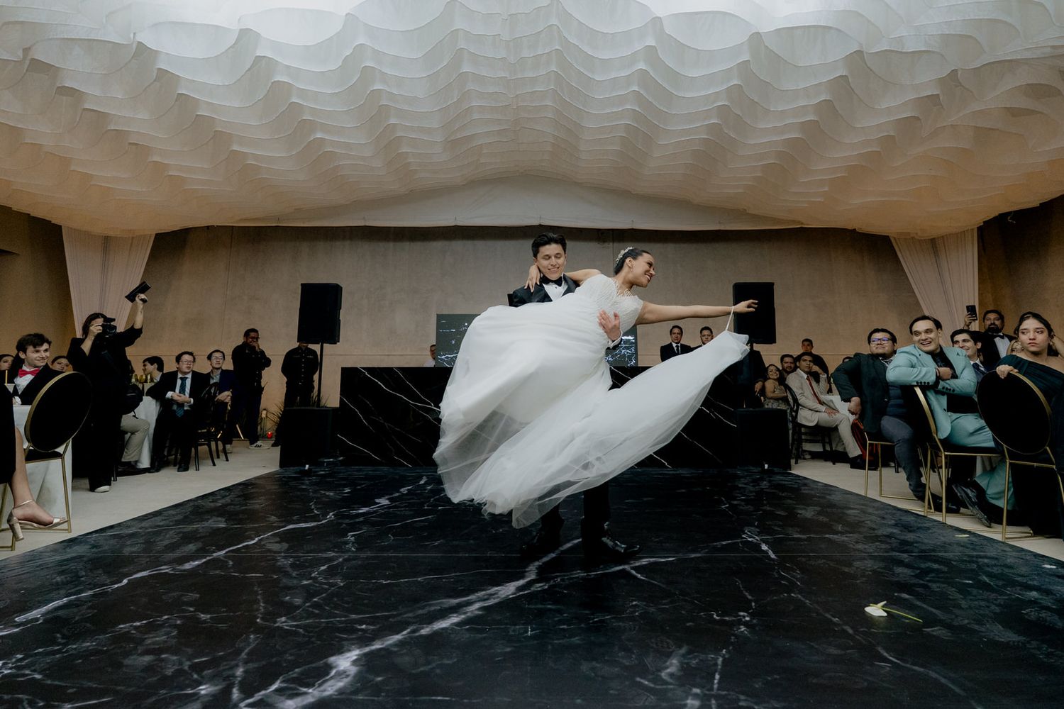 Groom lifts bride in flowing white gown during first dance on stunning black marble dance floor.