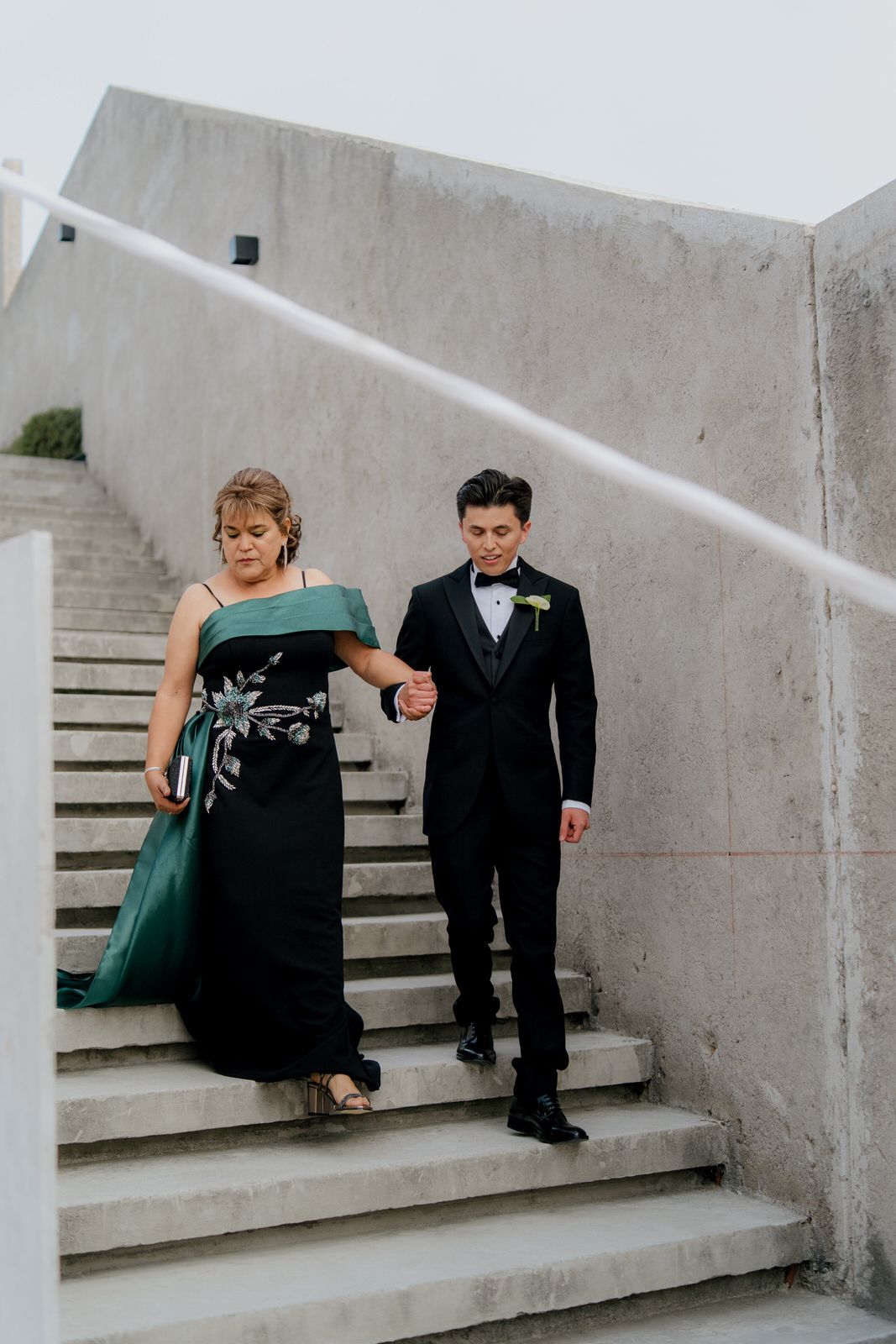 Groom in black tuxedo escorts elegantly dressed woman in green gown down concrete wedding venue stairs.