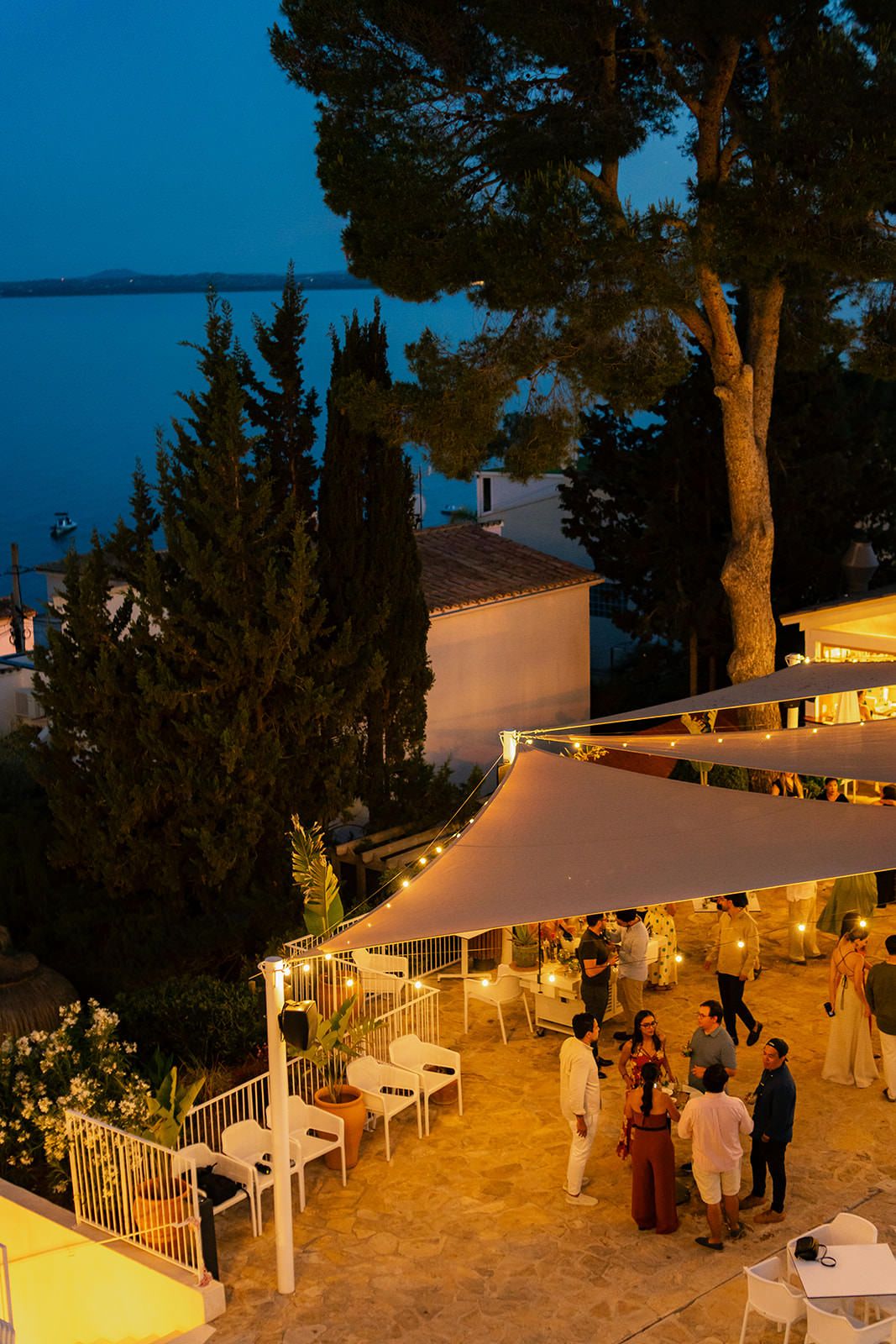 Nighttime view of an illuminated outdoor venue with guests gathering on a seaside terrace.