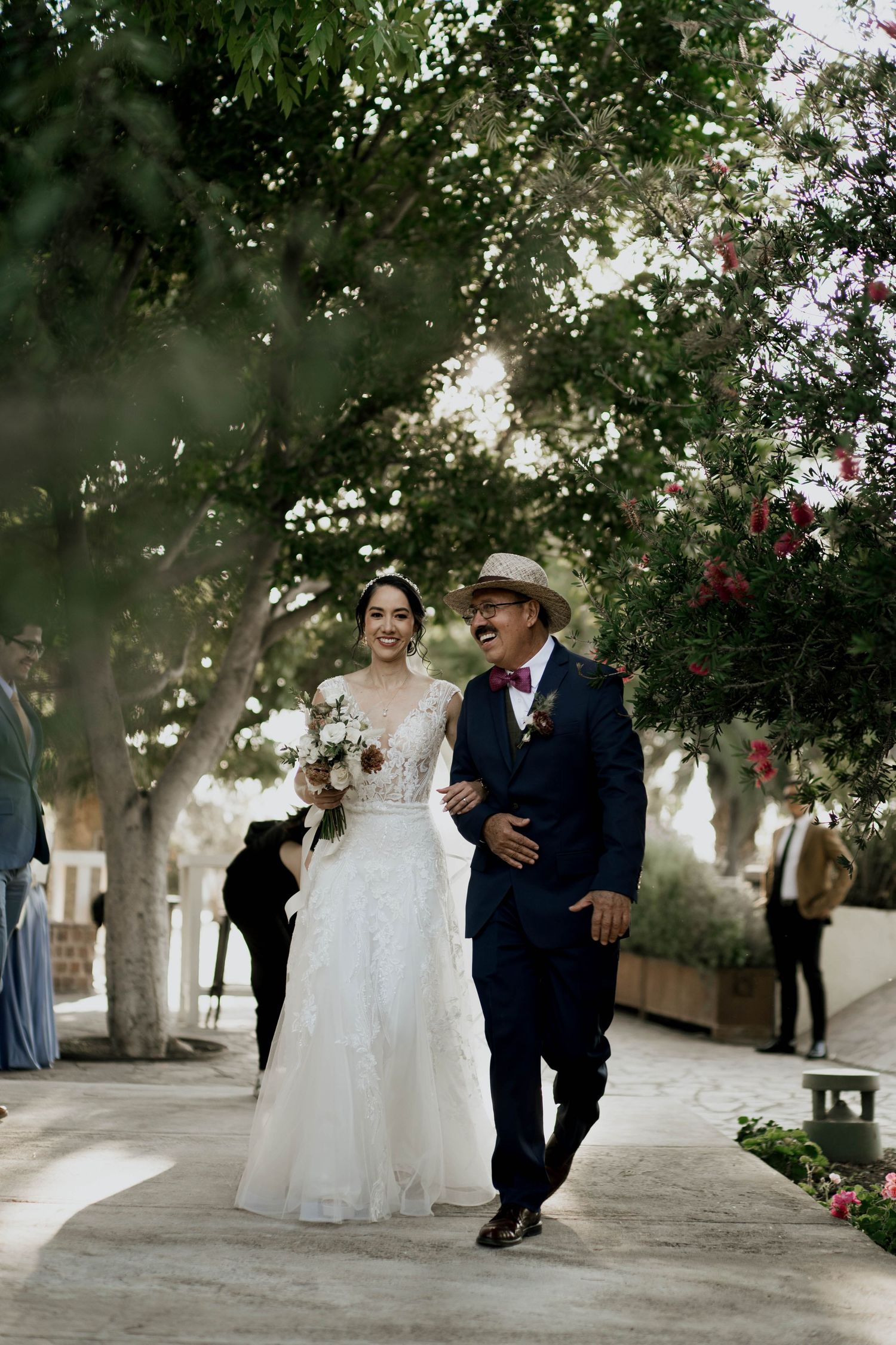 Couple strolls down tree-lined pathway during outdoor wedding celebration.