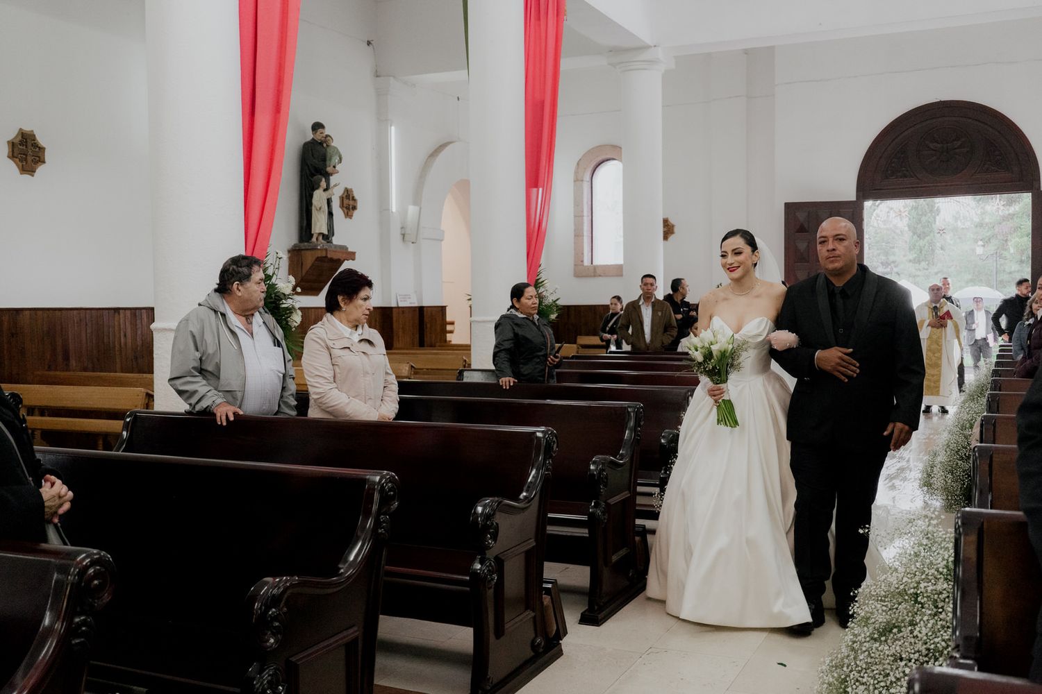 Wedding ceremony procession in a white church interior with red drapes and wooden pews.