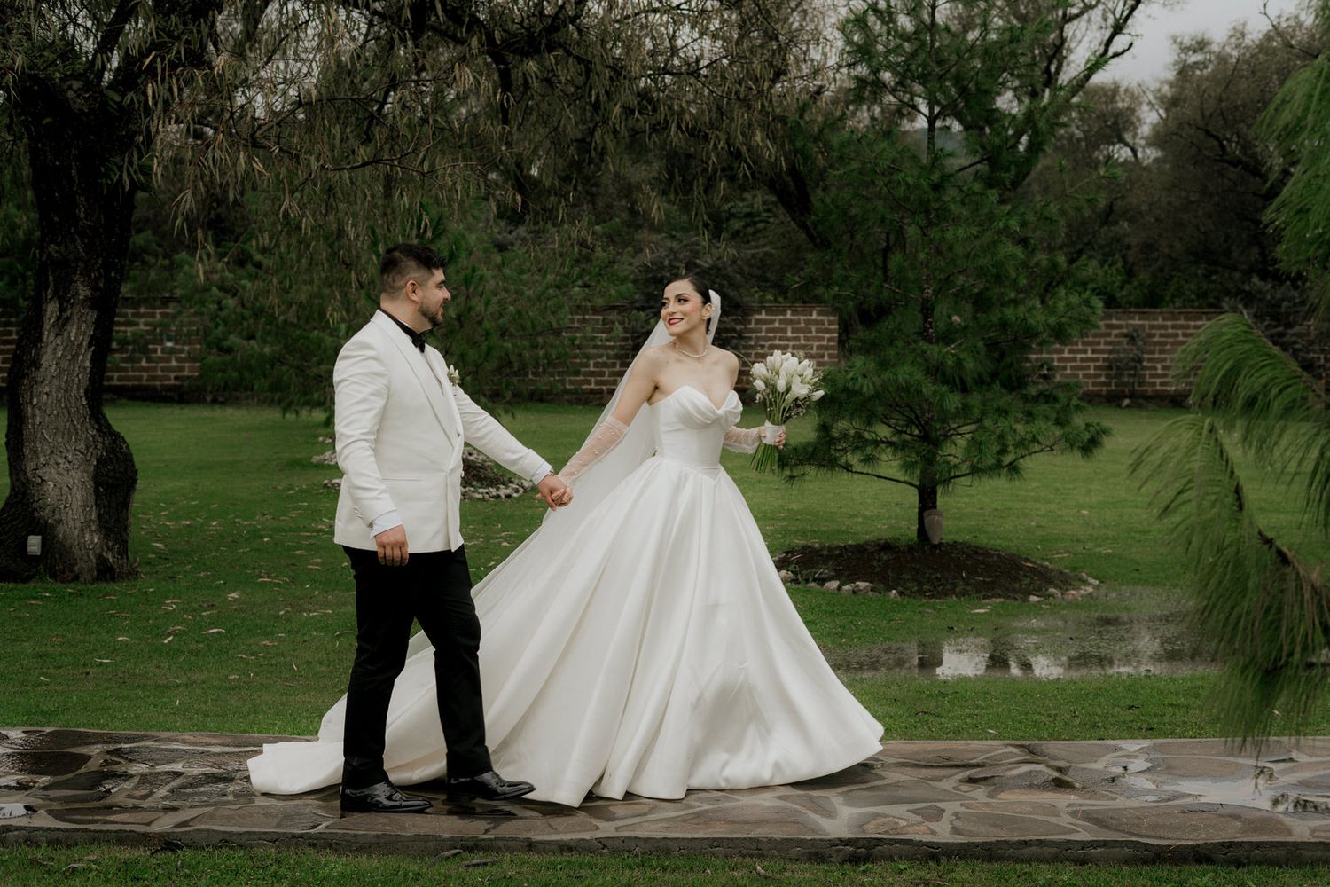 Couple in white wedding attire poses elegantly in lush garden setting.