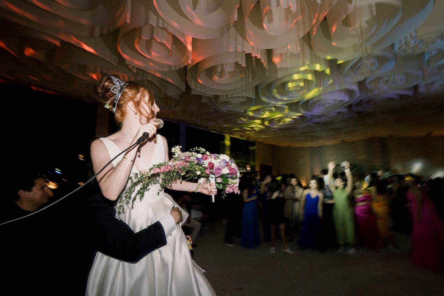 A bride in white holds a bouquet while dancing at a reception with colorful lighting effects on the ceiling.