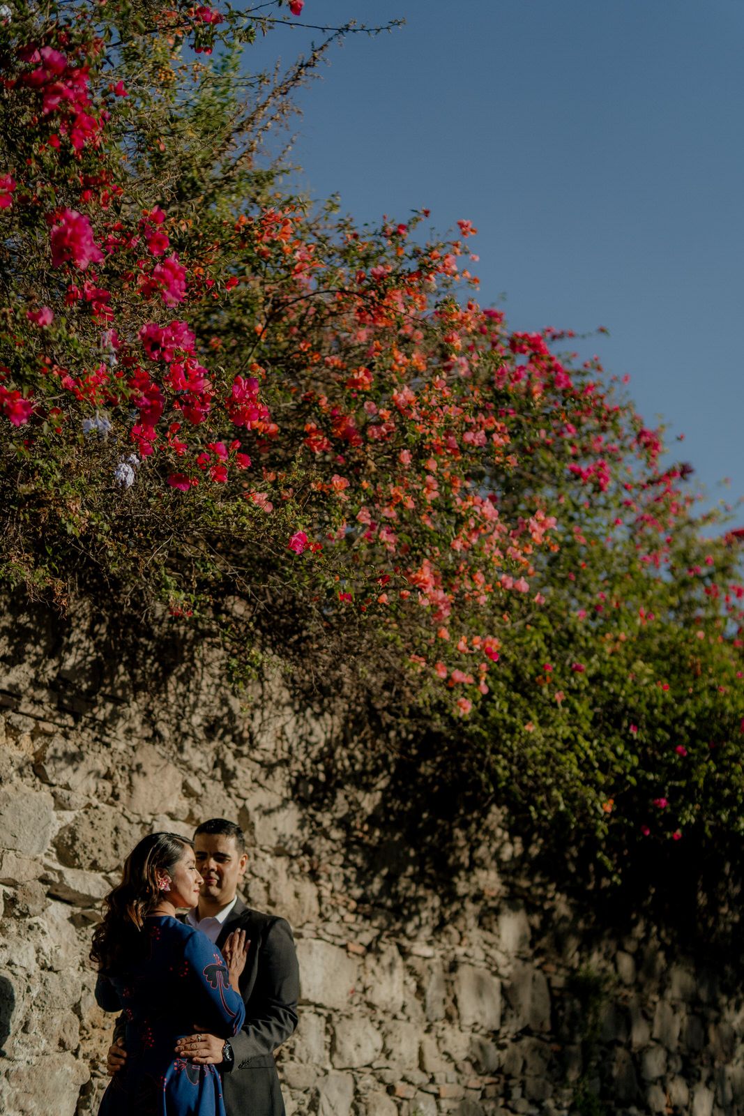 Pink bougainvillea flowers cascading over stone wall under bright blue sky.