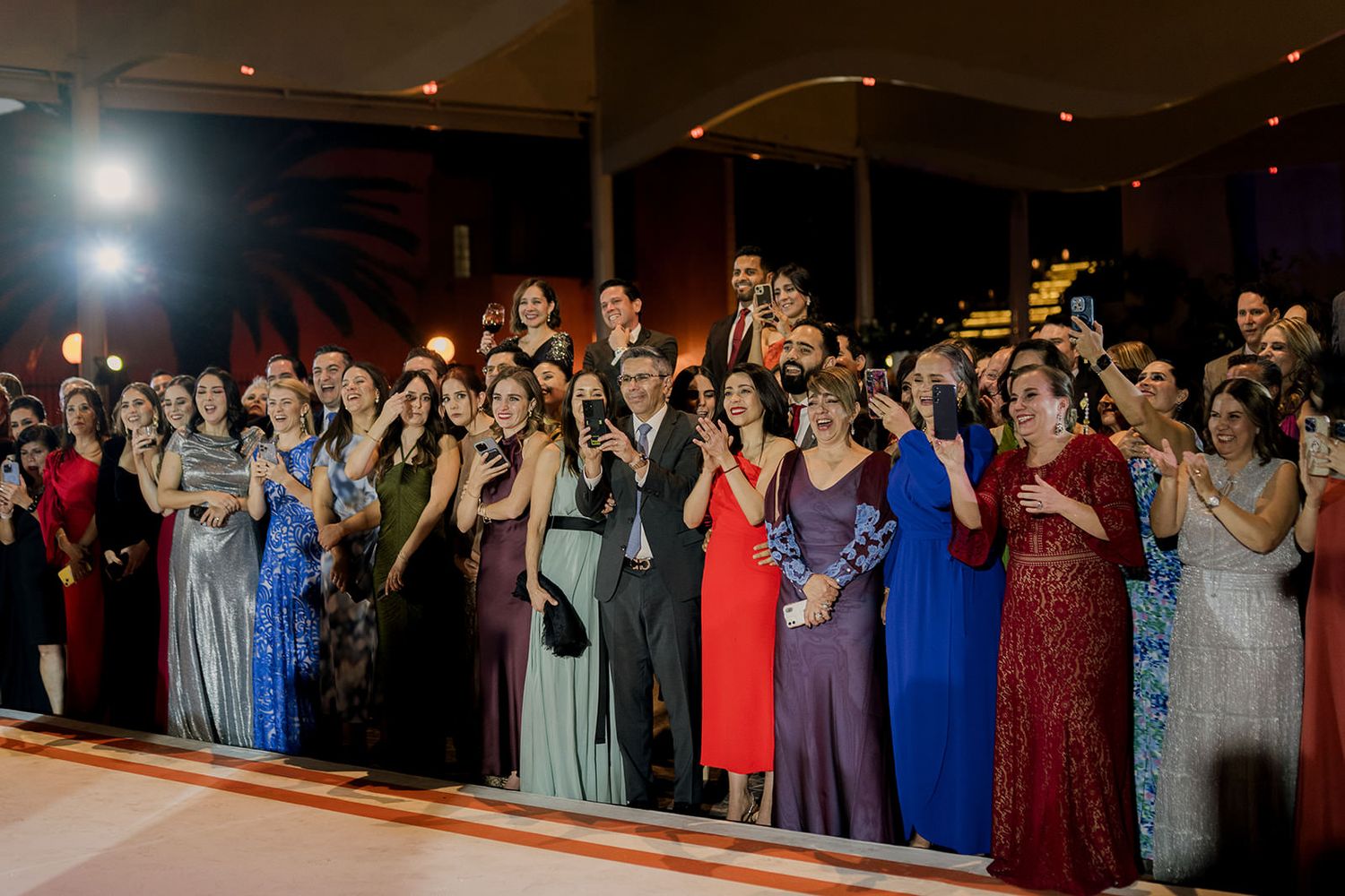 Elegantly dressed wedding guests in gowns applaud and celebrate at an indoor reception.