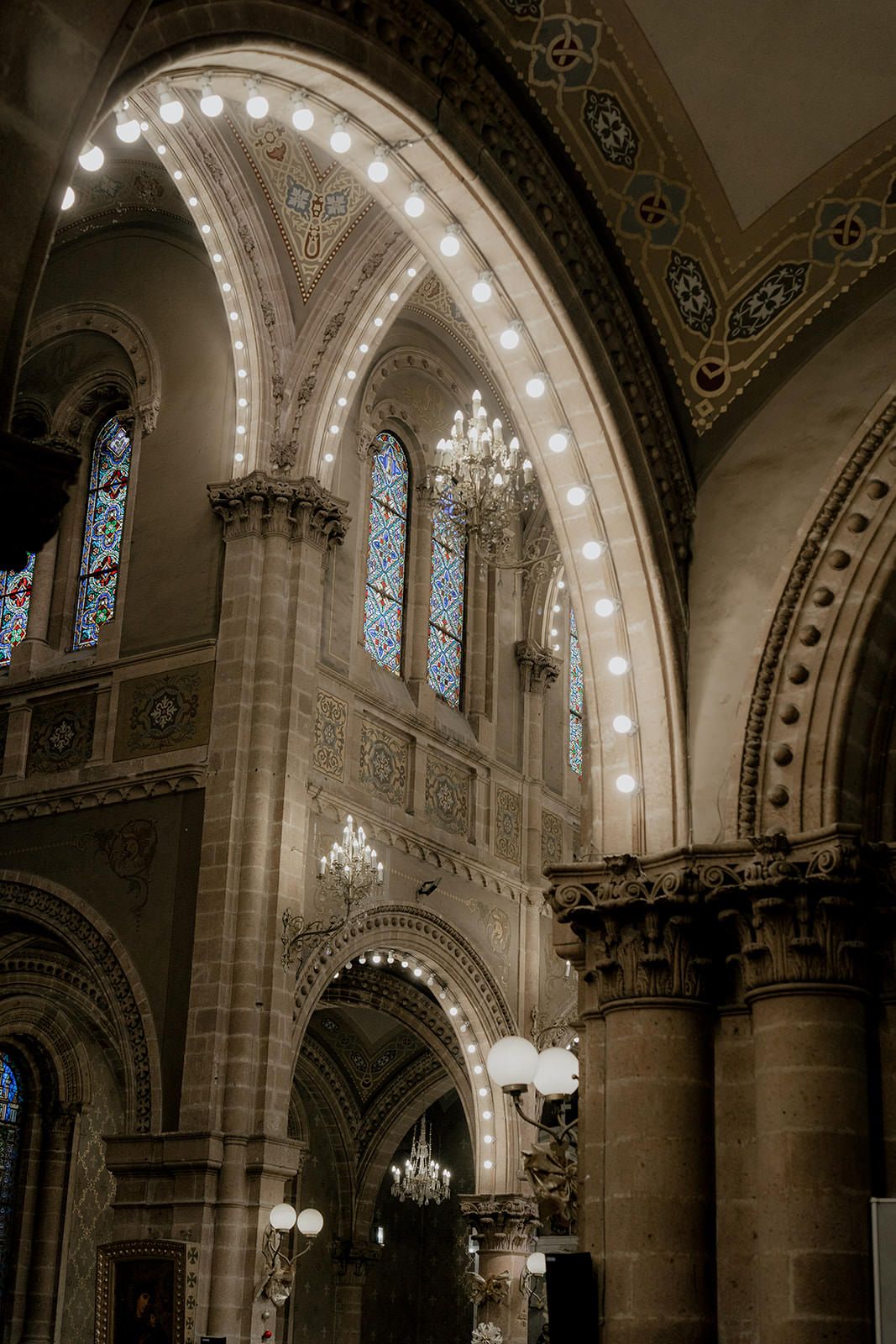 Gothic cathedral interior with illuminated arches, stained glass windows, ornate columns, and elegant chandeliers in sepia tone.