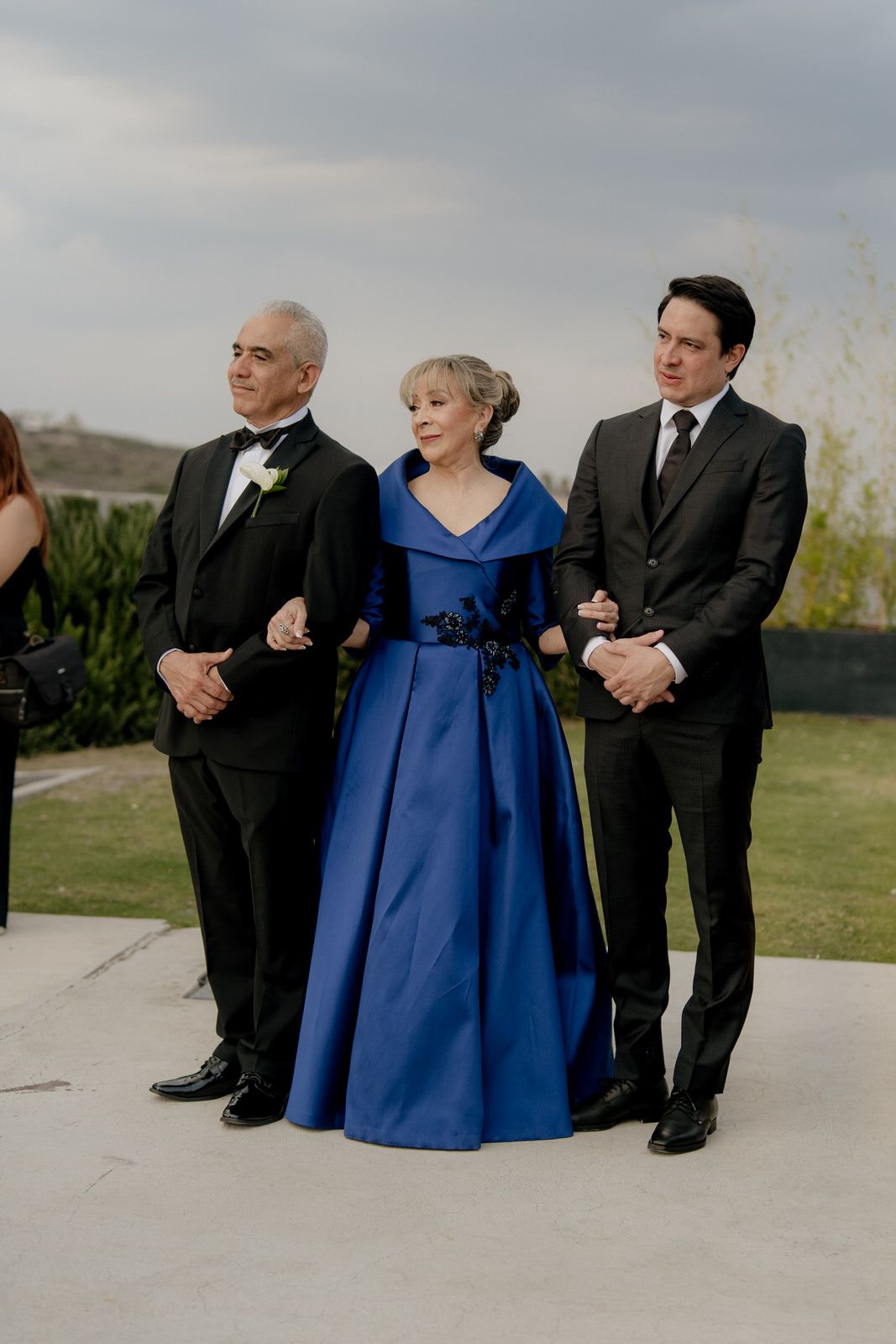 Elegant trio at formal event, woman in royal blue gown flanked by two men in black tuxedos outdoors.