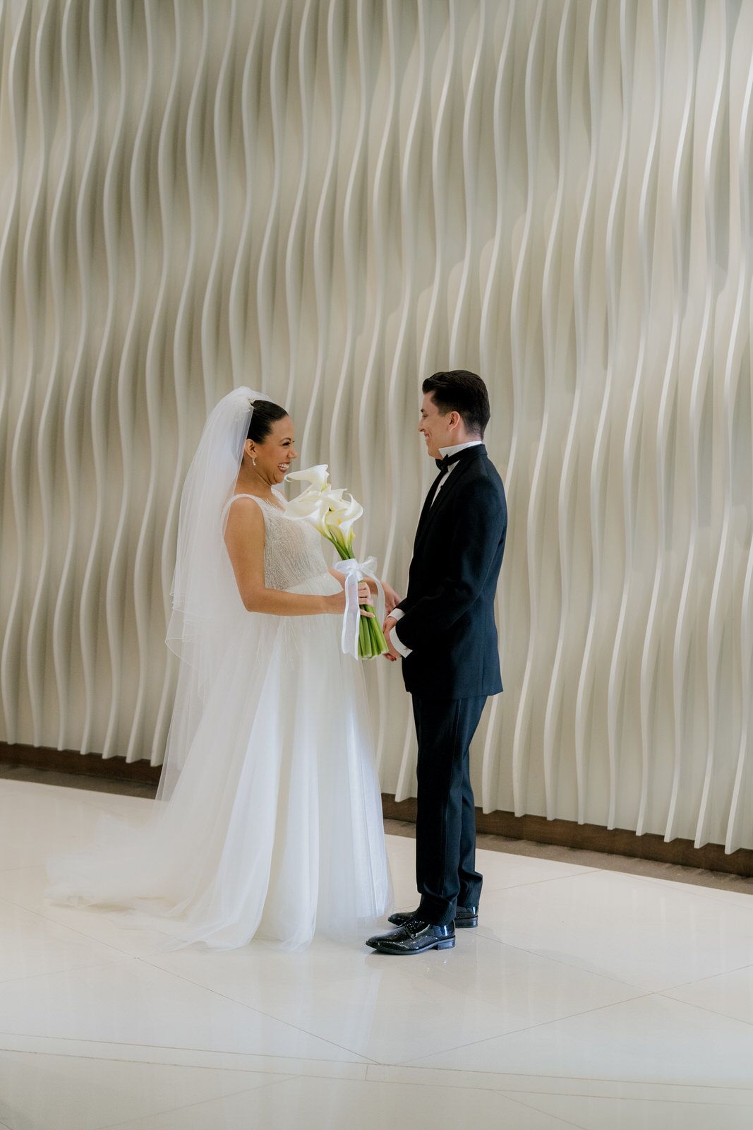 Bride in white gown holding calla lily bouquet exchanges vows with groom in black tuxedo before elegant wavy wall.
