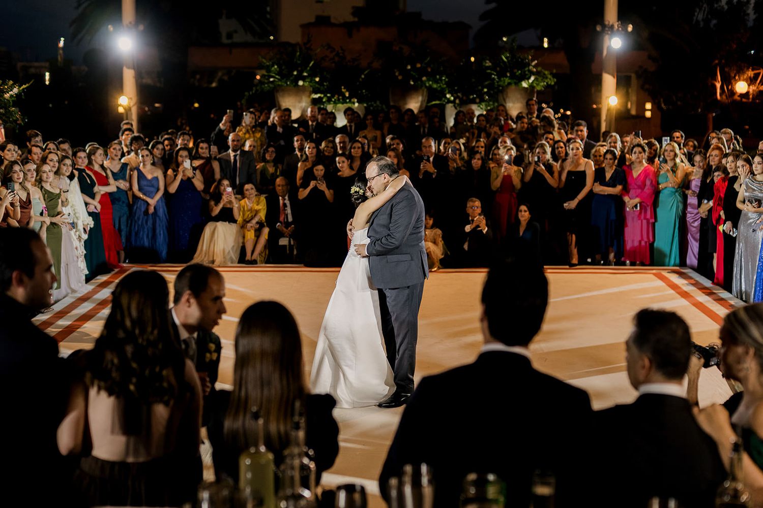 Newlyweds share romantic first dance on decorated floor surrounded by wedding guests at night.