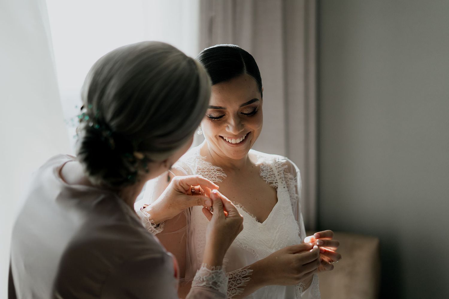 Smiling bride in lace wedding dress getting jewelry help from bridesmaid during wedding preparations.